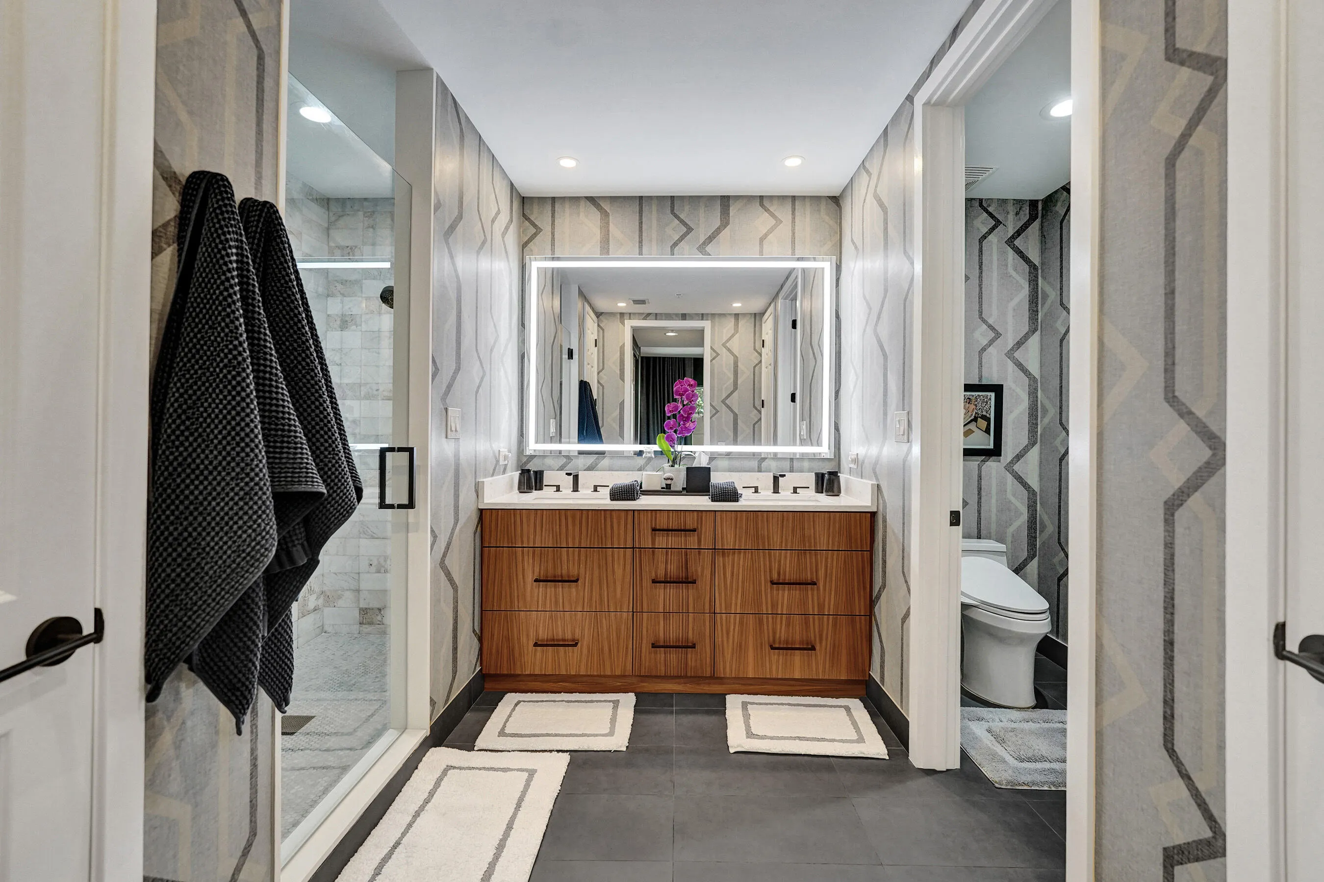 Double vanity with wood-finish cabinetry, illuminated mirror, walk-in shower with glass enclosure, and patterned wall covering