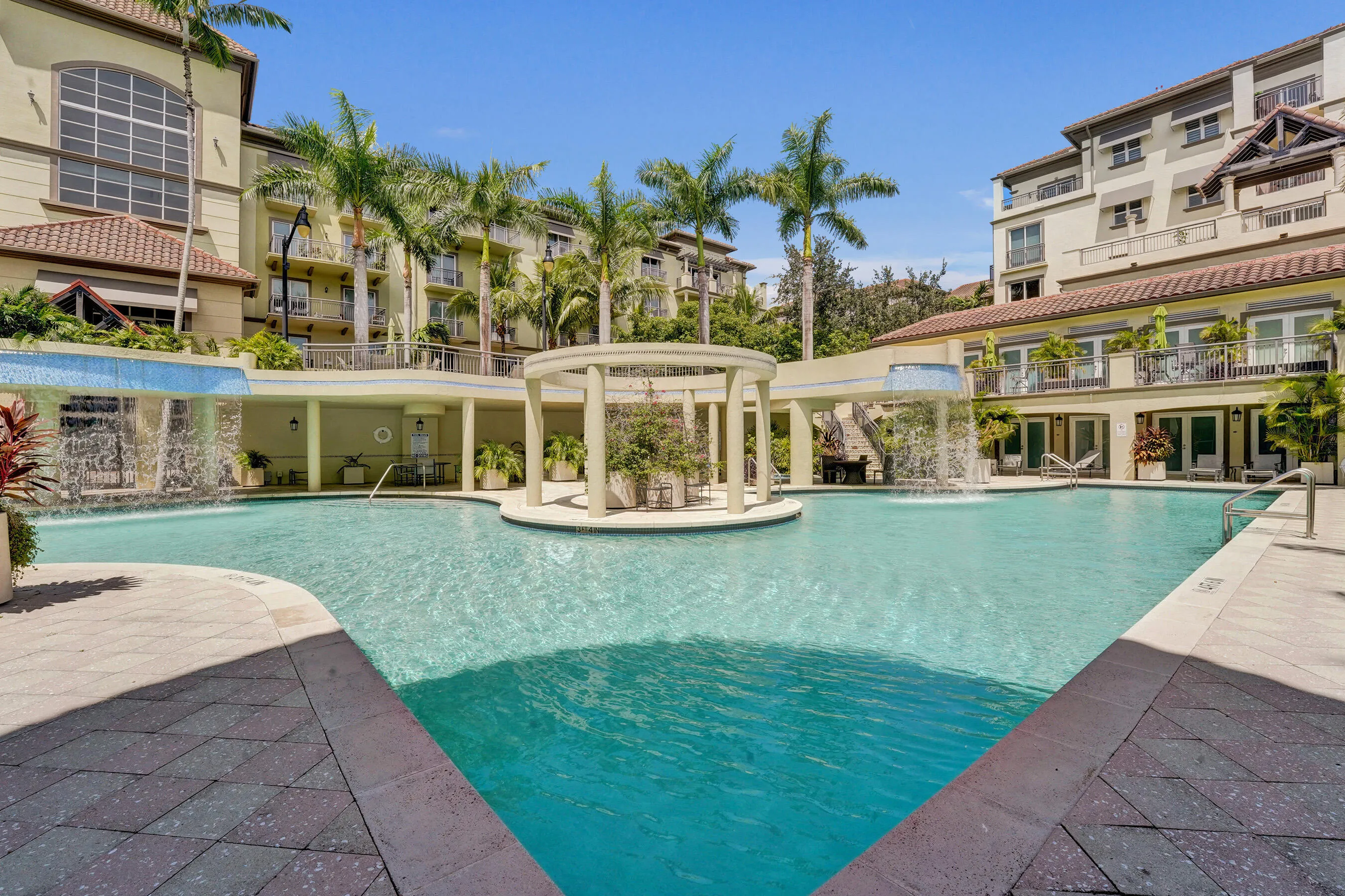 Expansive swimming pool featuring multiple water features, surrounding patio with paver stone, mature palm trees, and an architectural gazebo