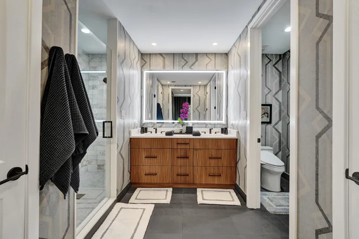 Double vanity with wood-finish cabinetry, illuminated mirror, walk-in shower with glass enclosure, and patterned wall covering