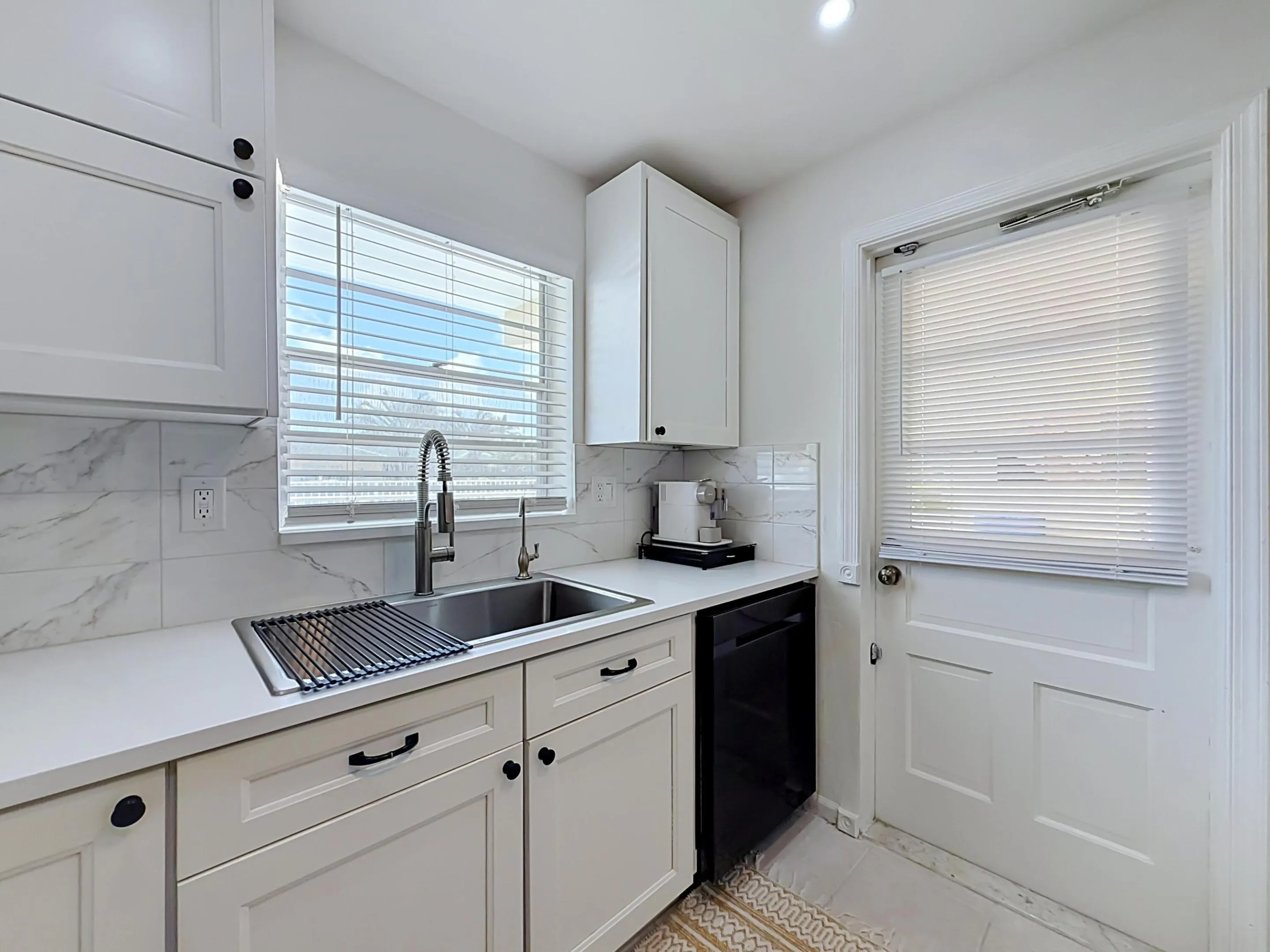 White shaker-style cabinetry with black hardware, undermount stainless steel sink, pull-down faucet, white solid surface countertops, and marble-look tile backsplash