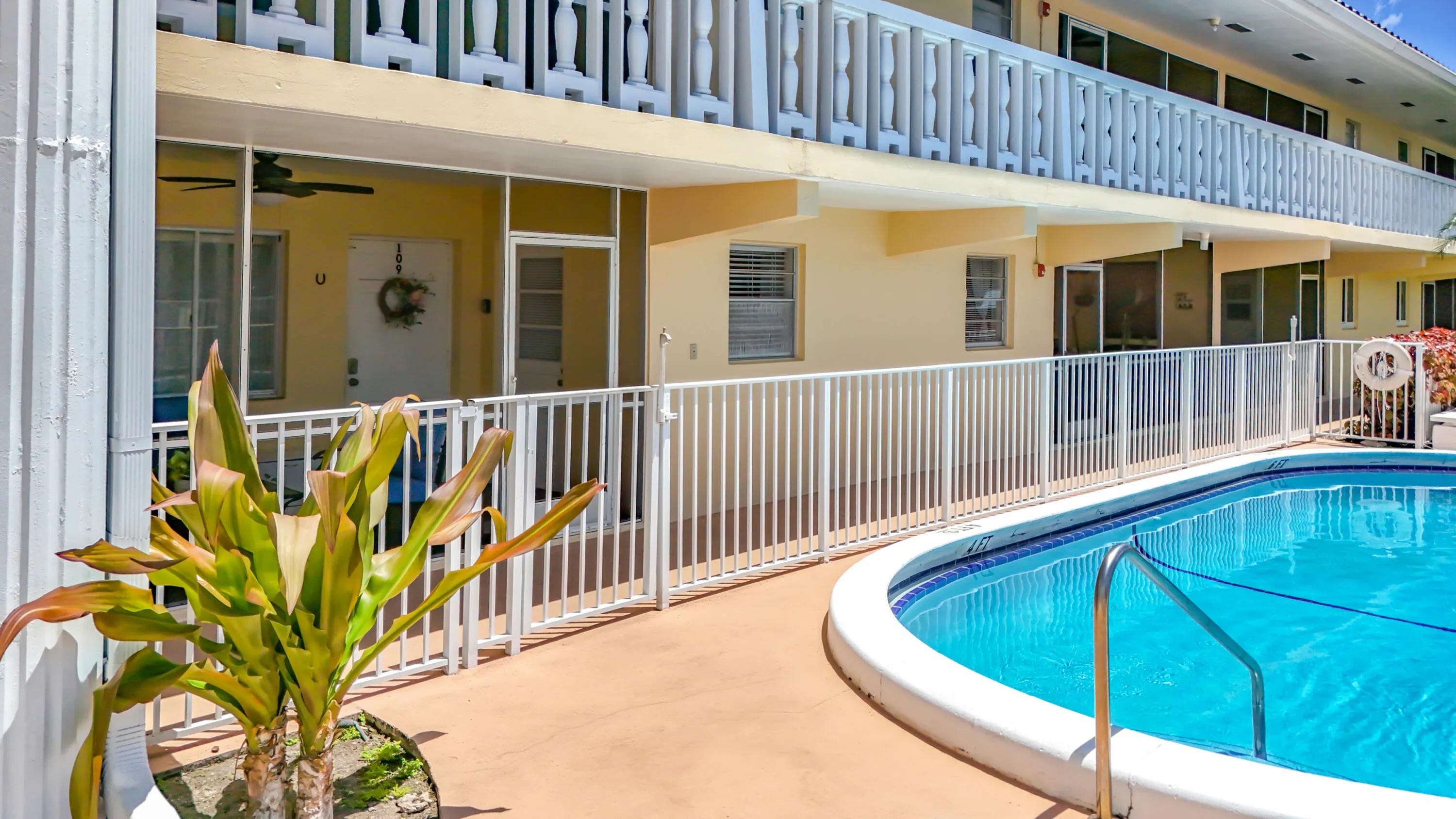 Curved swimming pool with white coping, surrounding paved deck, and adjacent white fence
