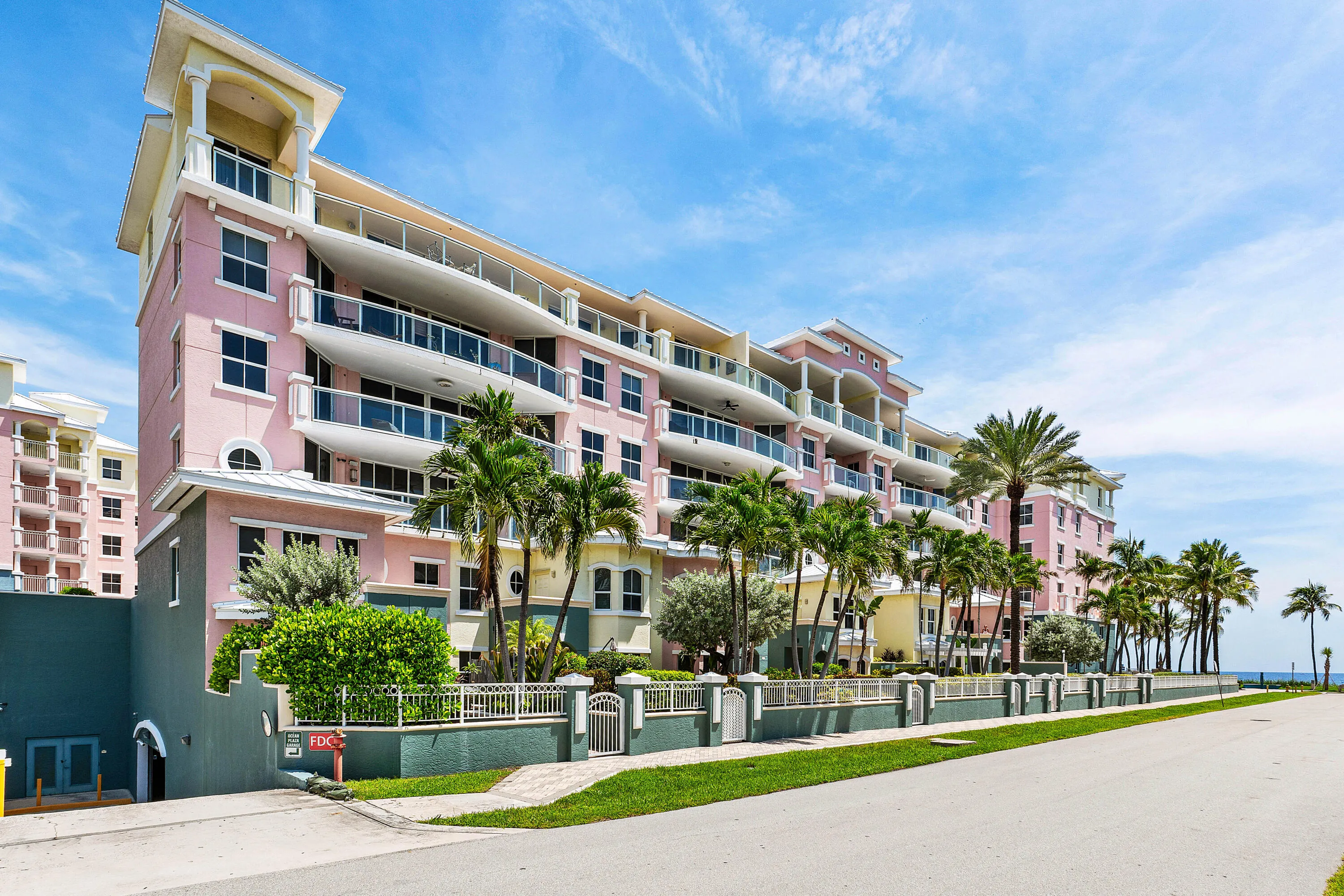 Multi-story condominium building featuring private balconies, arched window details, a decorative exterior fence, and extensive landscaping with palm trees