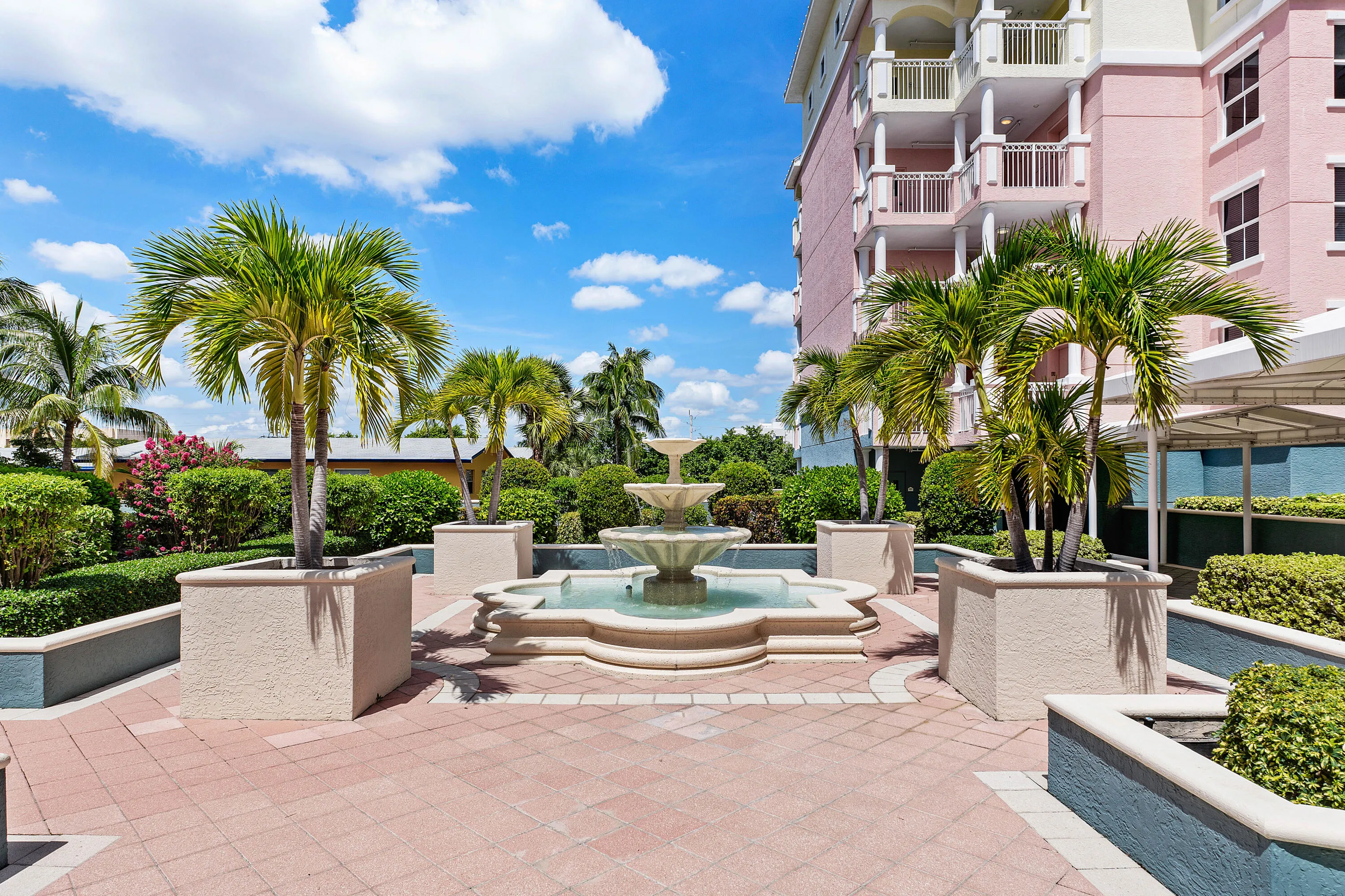 Three-tiered fountain, paver patio, palm trees in planters, manicured hedges, and multi-story building exterior