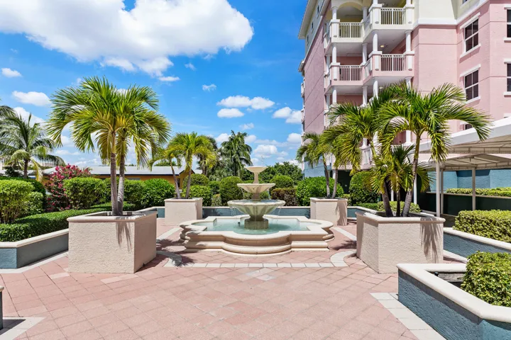 Three-tiered fountain, paver patio, palm trees in planters, manicured hedges, and multi-story building exterior