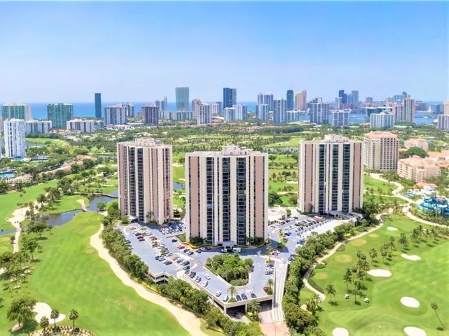 Three high-rise buildings situated amidst a golf course landscape with a distant city skyline and ocean background