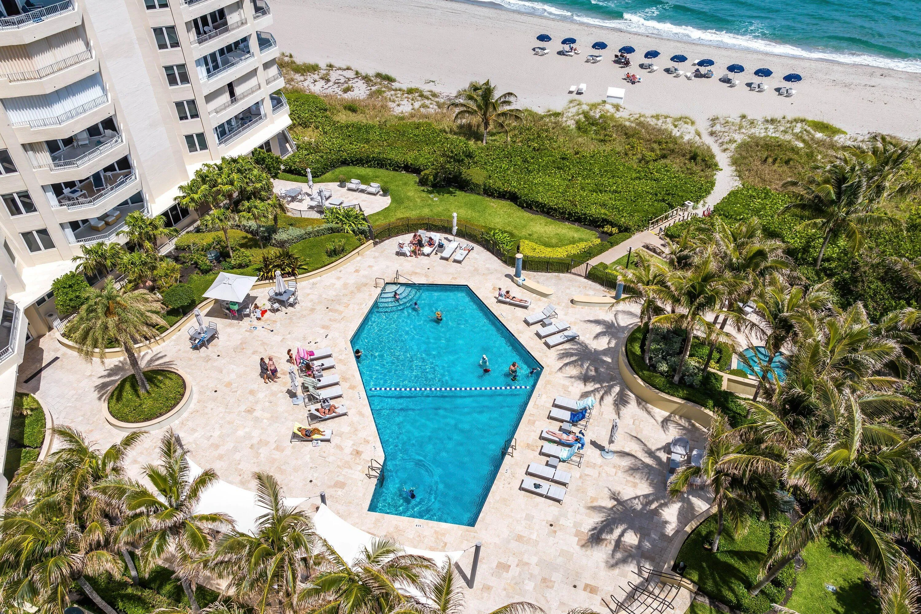 Geometric swimming pool with surrounding paver deck, mature palm trees, and manicured green spaces bordering a sandy beach with ocean waves