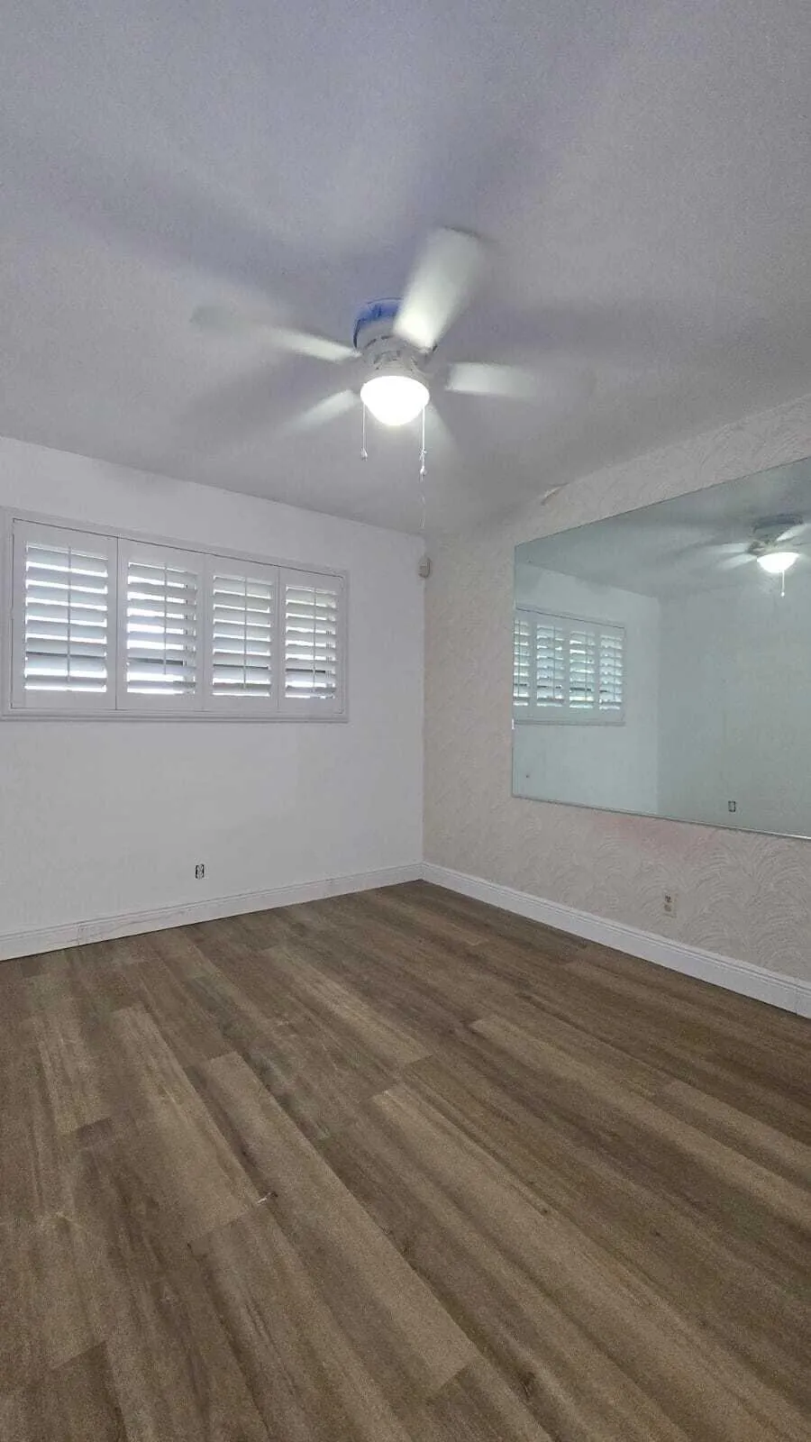 White walls, wood-finish flooring, ceiling fan with integrated lighting, window with white plantation shutters, and a large wall-mounted mirror
