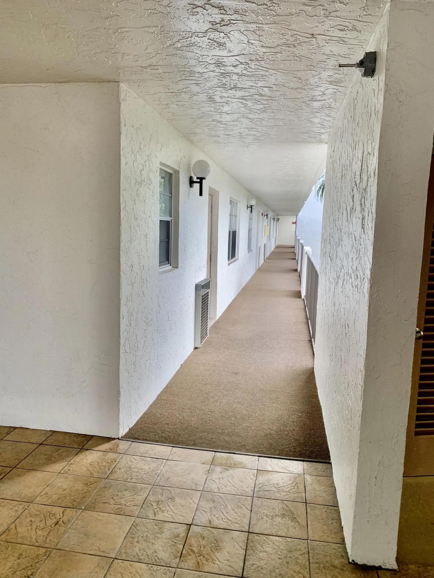Covered exterior walkway featuring textured wall finishes, sconce lighting fixtures, and a carpeted path