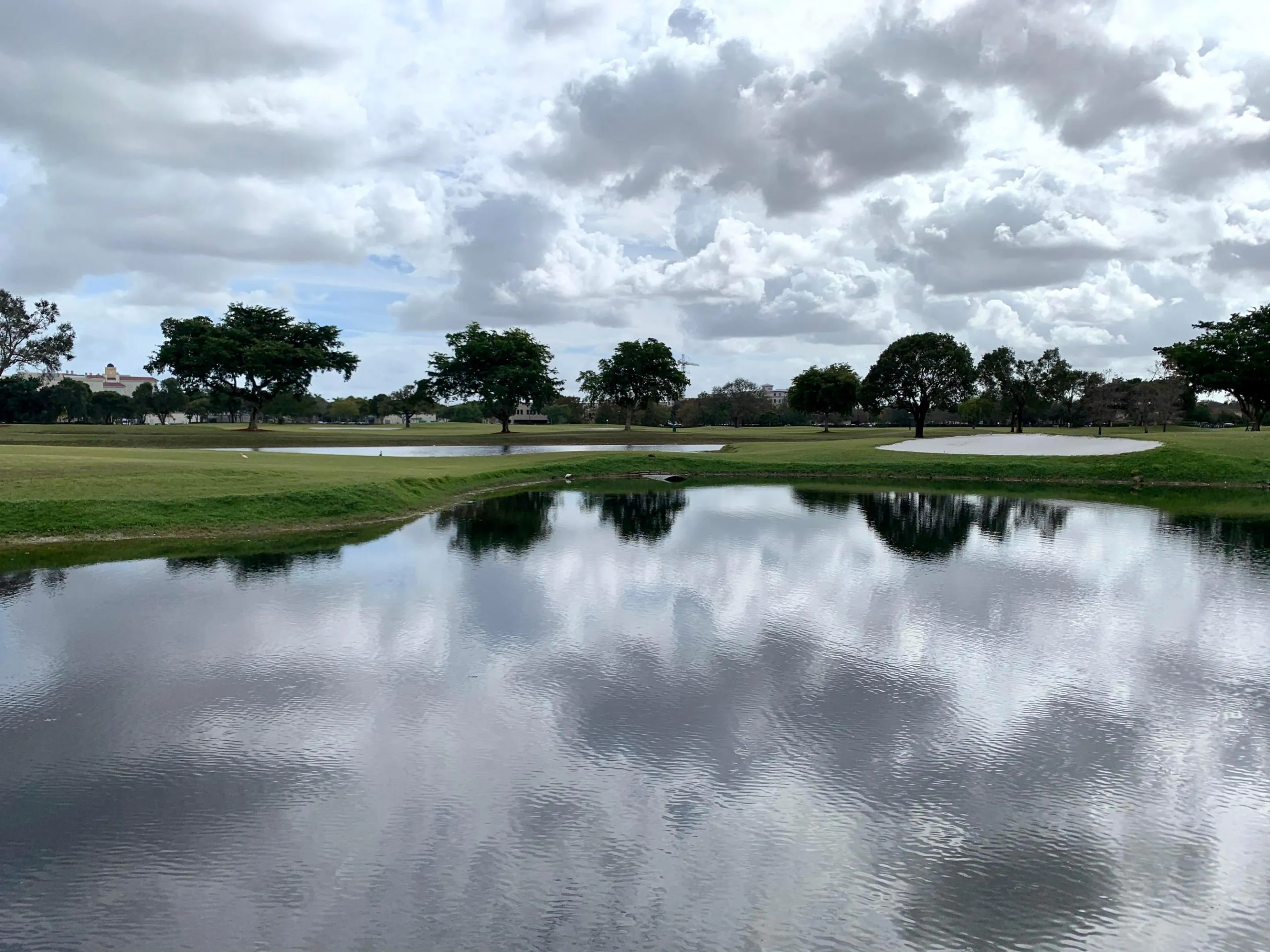 Expansive golf course frontage featuring a water hazard, manicured green space, mature trees, and a distant sand trap