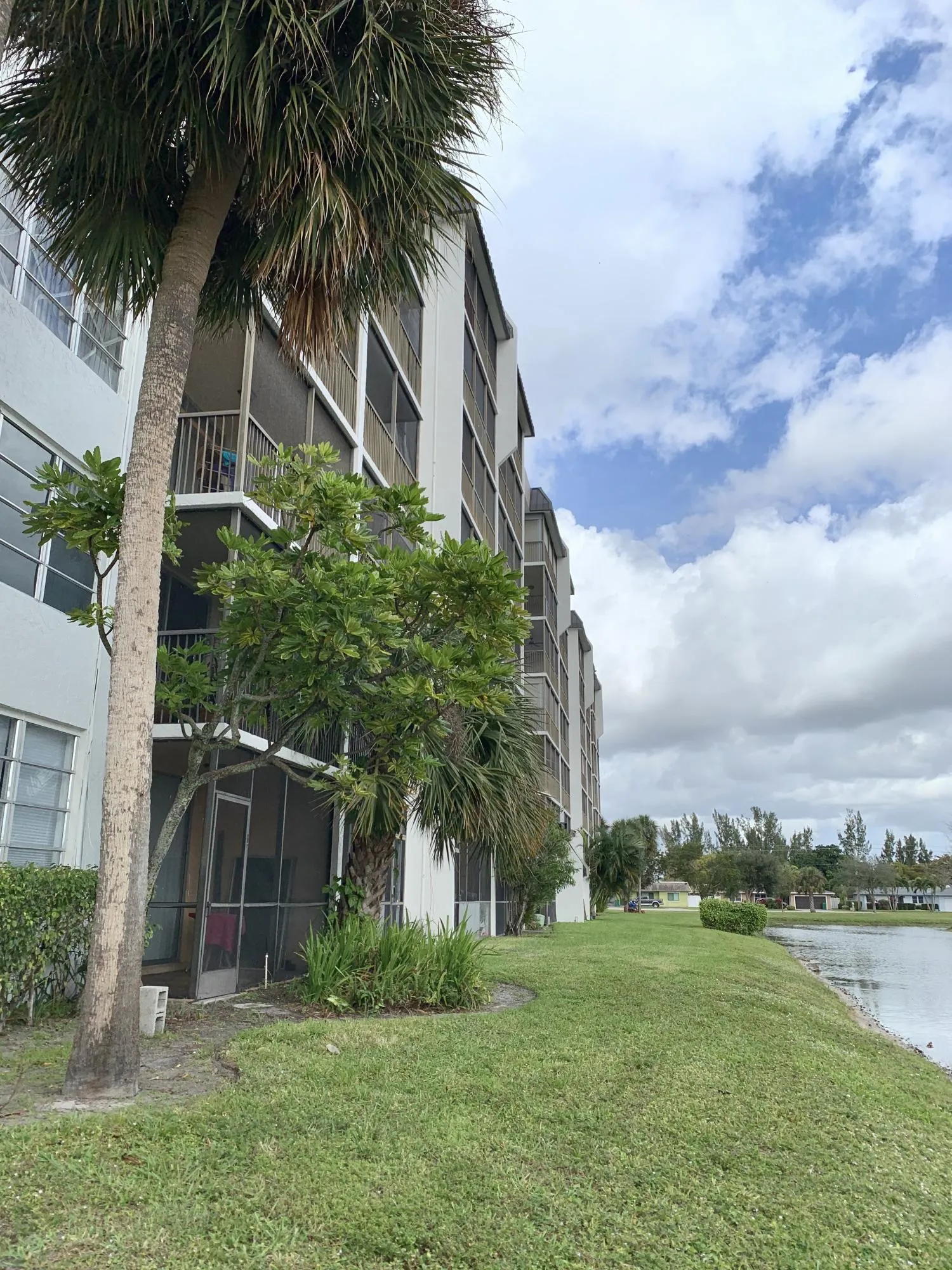 Multi-story building featuring screened balconies and patios, mature palm trees, manicured lawn, and waterside frontage