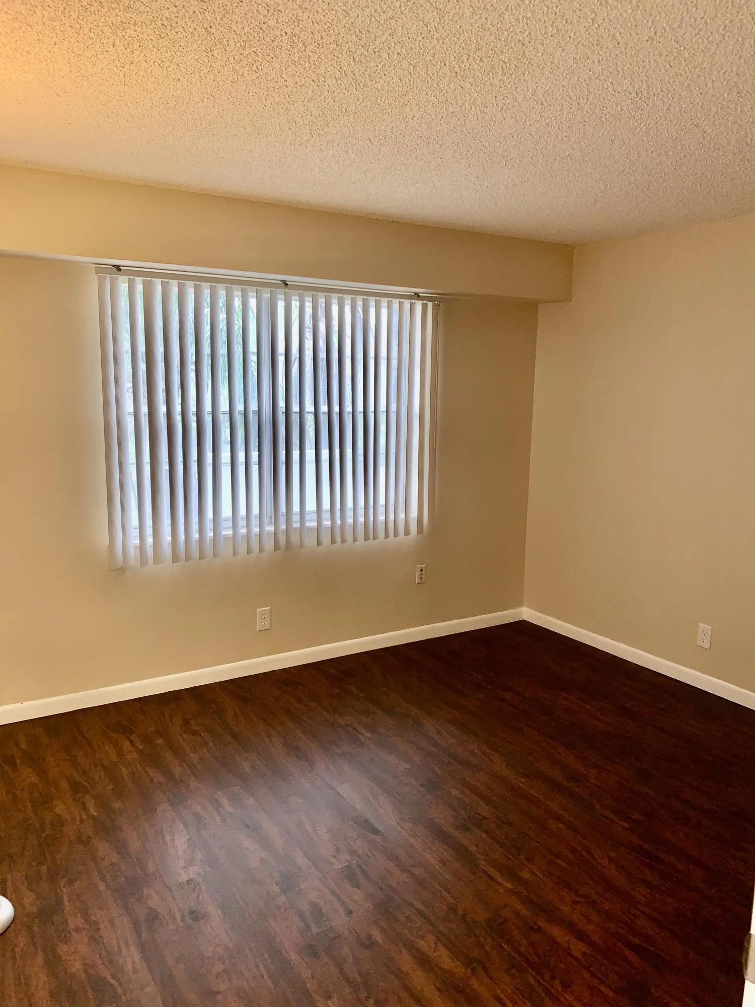 Spacious room featuring rich wood-finish flooring, neutral wall tones, and a large window with vertical blinds