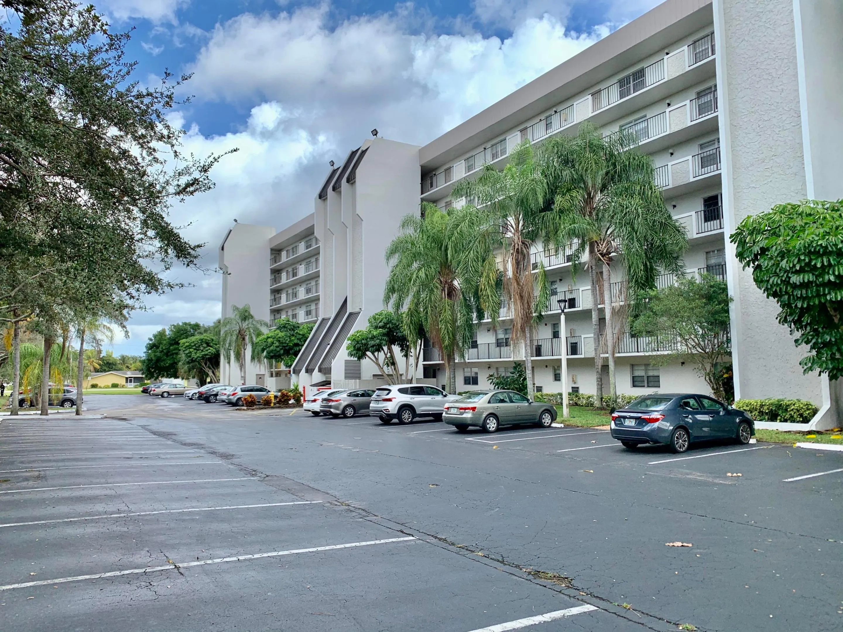 Multi-story residential building featuring private balconies with white railings, mature palm trees, and ample paved parking