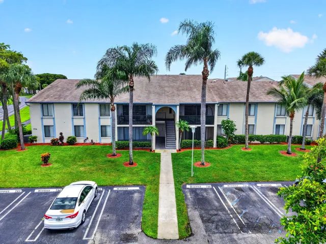 Two-story residential building with light-colored exterior and blue trim accents, featuring multiple screened balconies, a central exterior staircase, and a paved parking area
