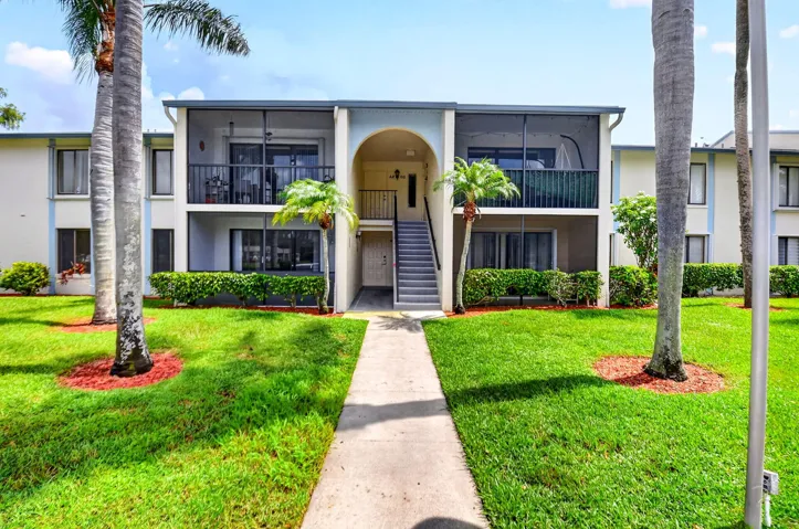 Two-story building with screened balconies, mature palm trees, and a concrete walkway