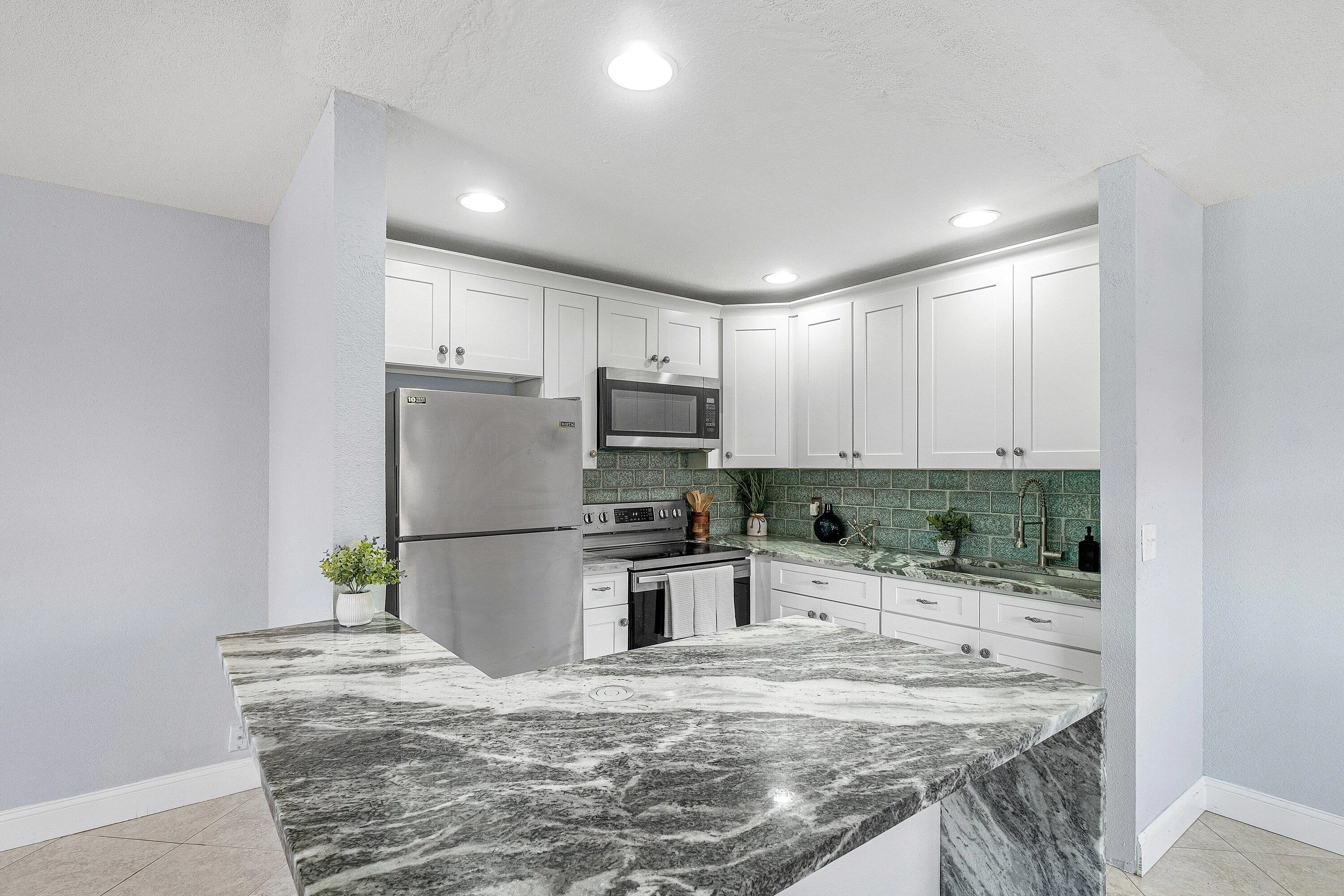 Kitchen featuring white shaker cabinetry, stainless steel appliances, a green subway tile backsplash, and a large countertop with a waterfall edge