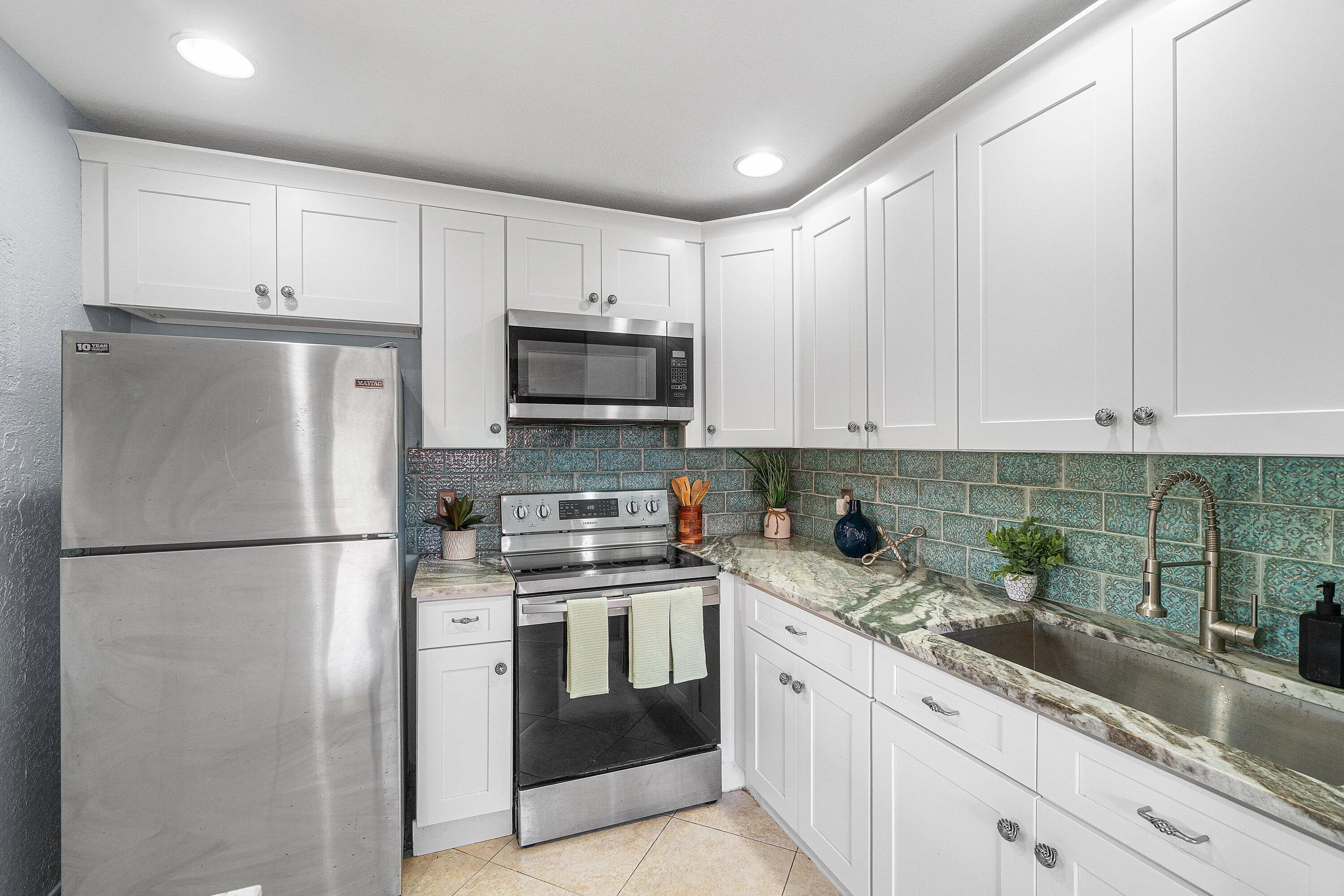 White shaker-style cabinetry, stainless steel appliances, variegated green backsplash, stone countertops, and a stainless steel sink with a gooseneck faucet