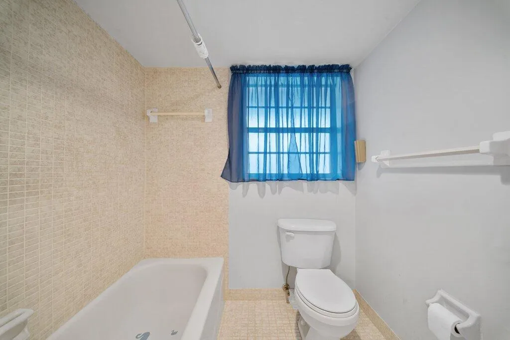 White bathtub with full-height tiled surround, white toilet, and window with privacy​​‌​​​​‌​​‌‌​‌‌​​​‌‌​‌​‌​‌​​​‌​​ grid