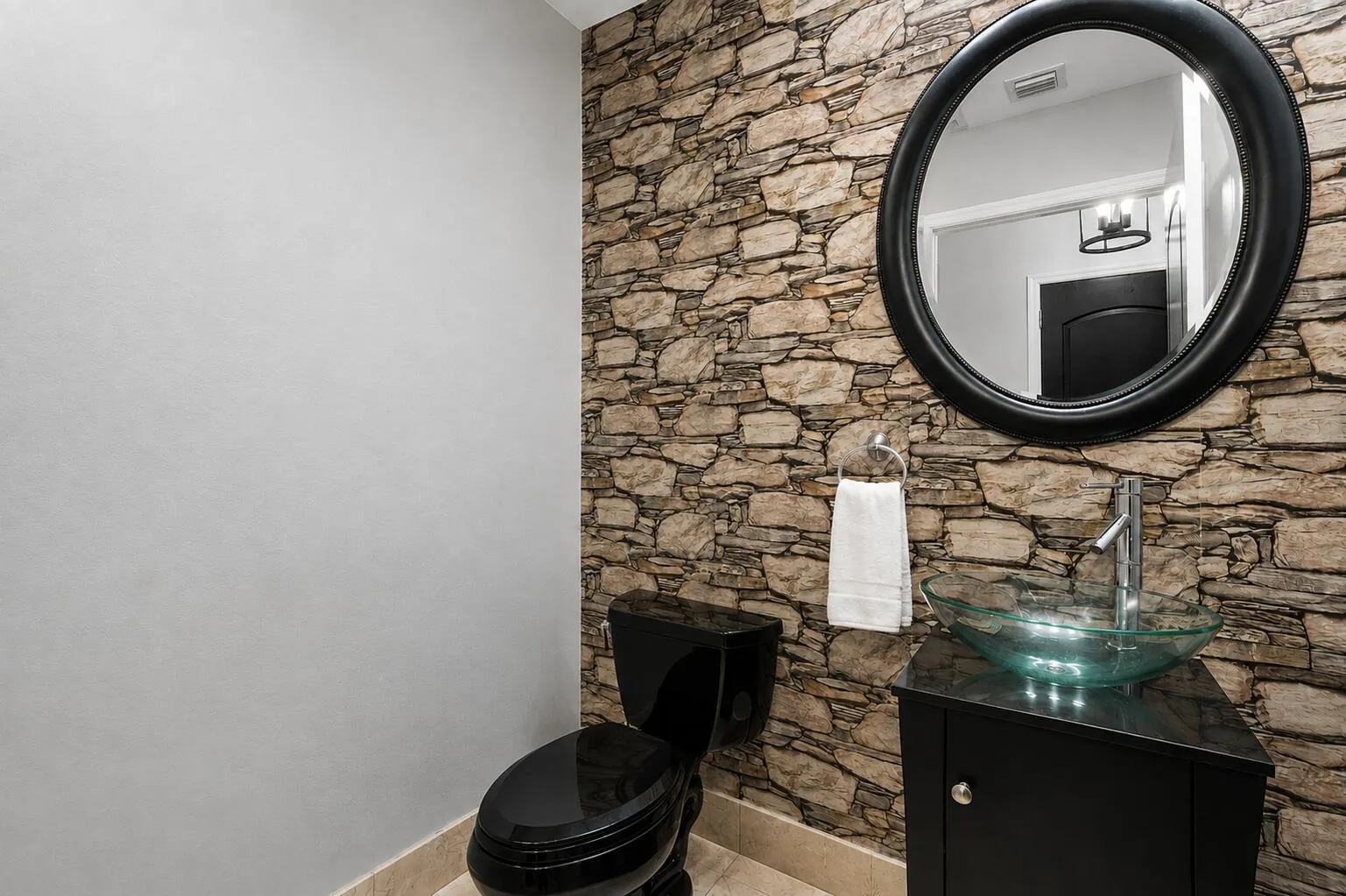 Stone-finish accent wall with a black framed round mirror, black vanity with a glass vessel sink, and a black toilet