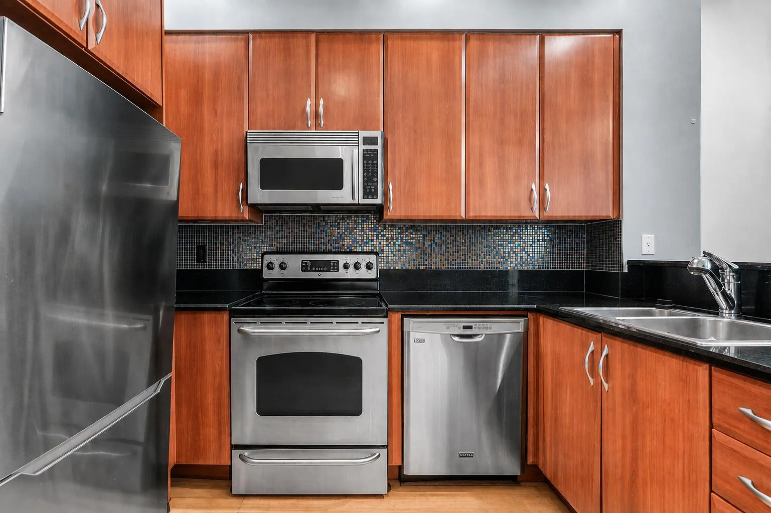 Kitchen featuring stainless steel appliances, wood-finish cabinetry, black countertops, and a mosaic tile backsplash