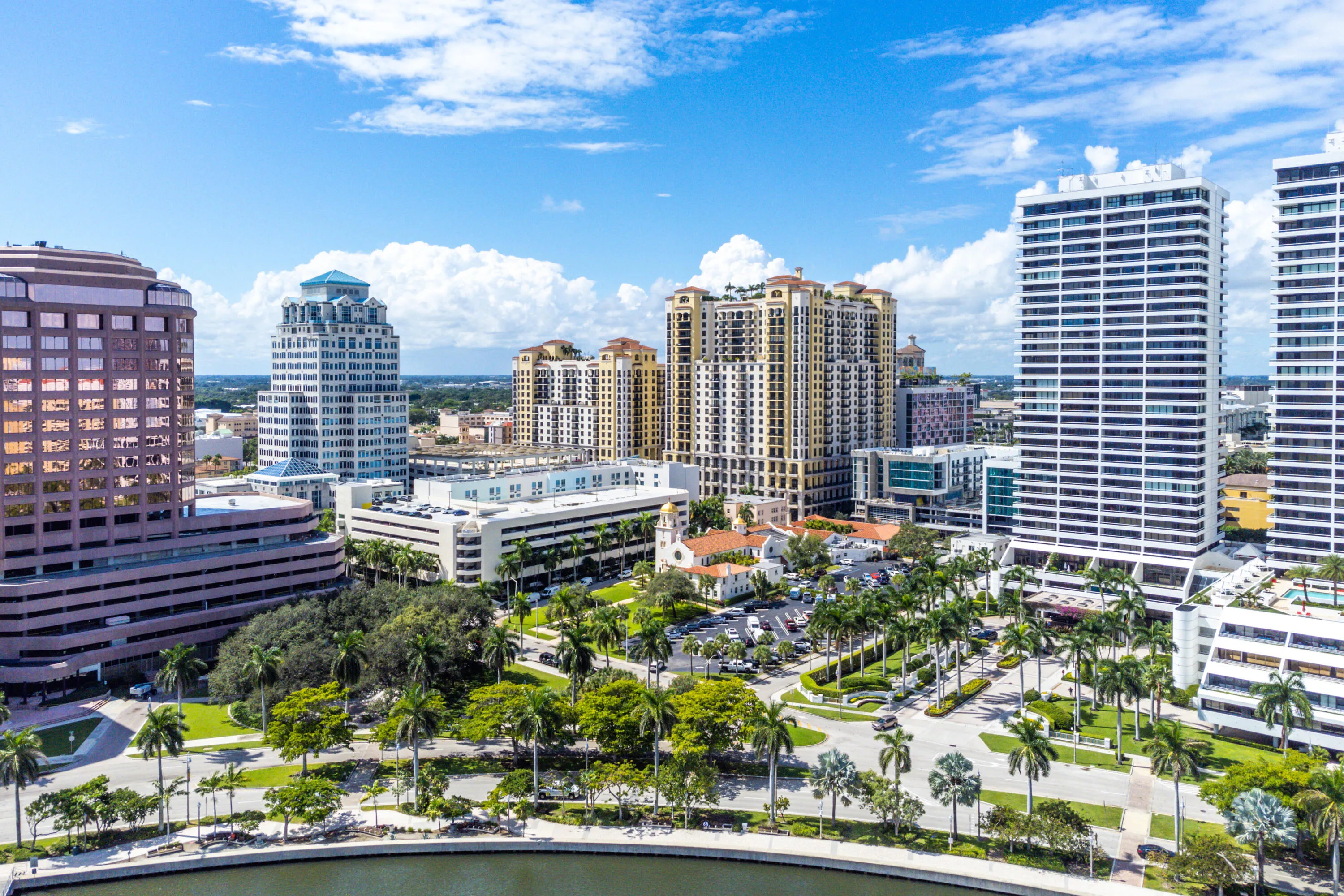 Urban landscape featuring multiple high-rise buildings, mature palm trees, and a prominent body of water