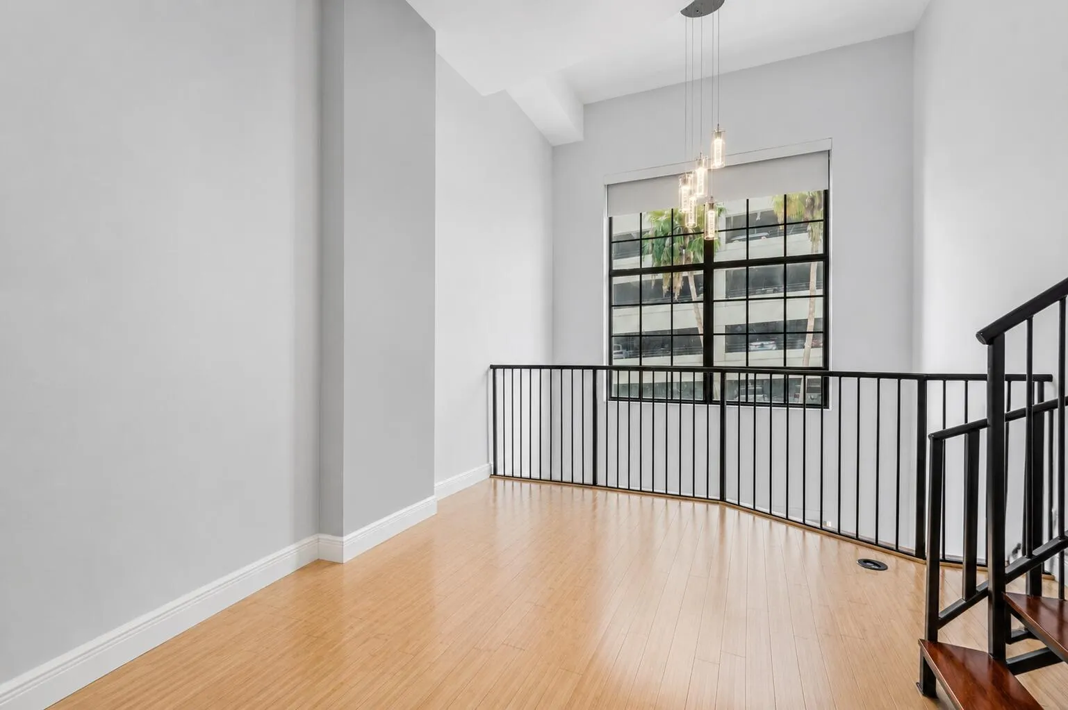 High ceilings with a suspended light fixture, large framed window with roller shade, wood-finish flooring, and a black wrought iron railing