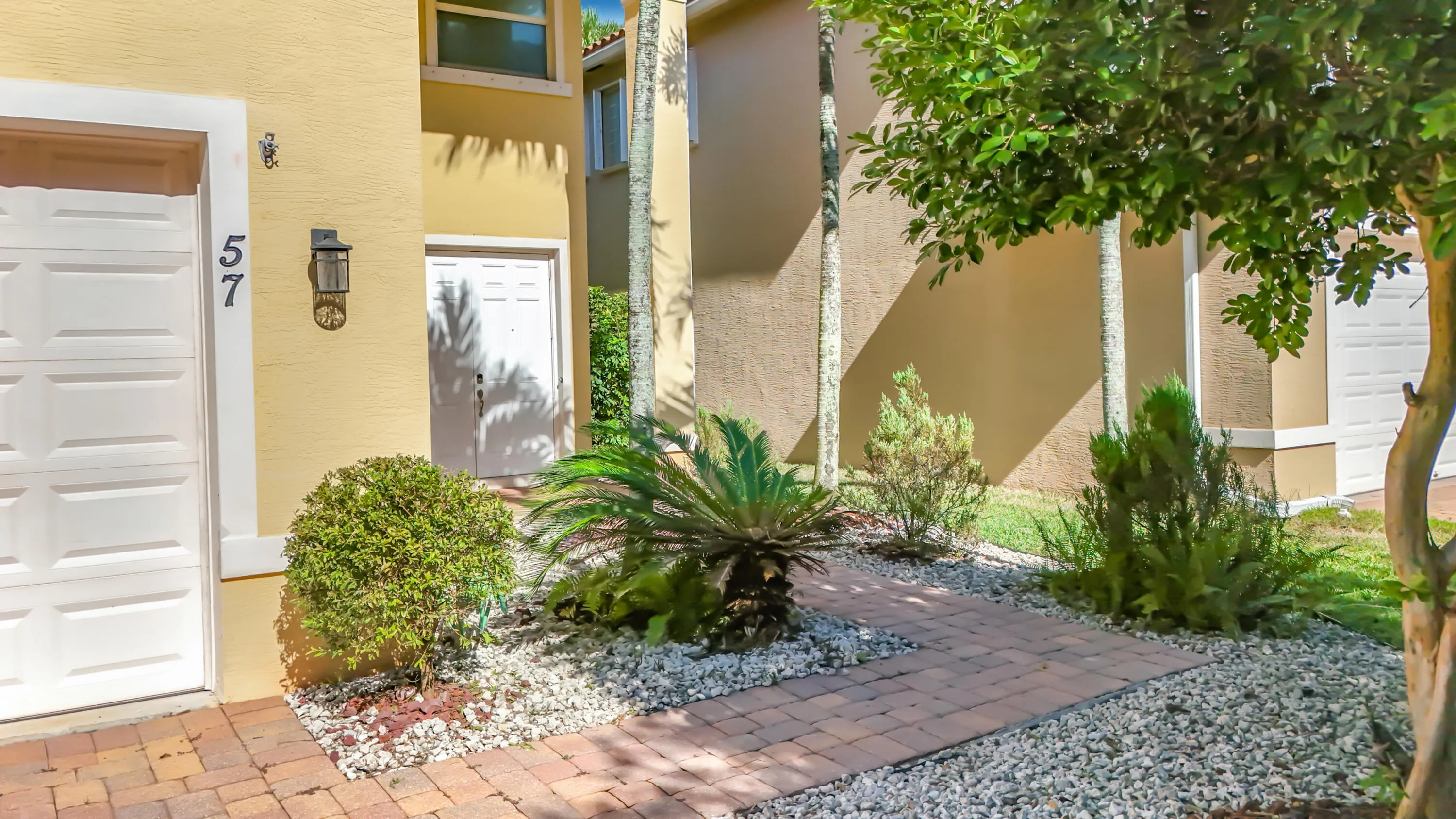 Exterior featuring a brick paver pathway, white garage door, exterior wall lantern, and manicured landscaping with white gravel