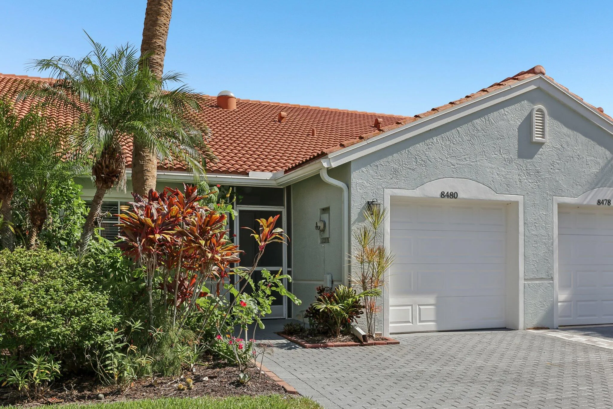 Stucco exterior with a terracotta tile roof, garage with a paved driveway, and mature tropical landscaping