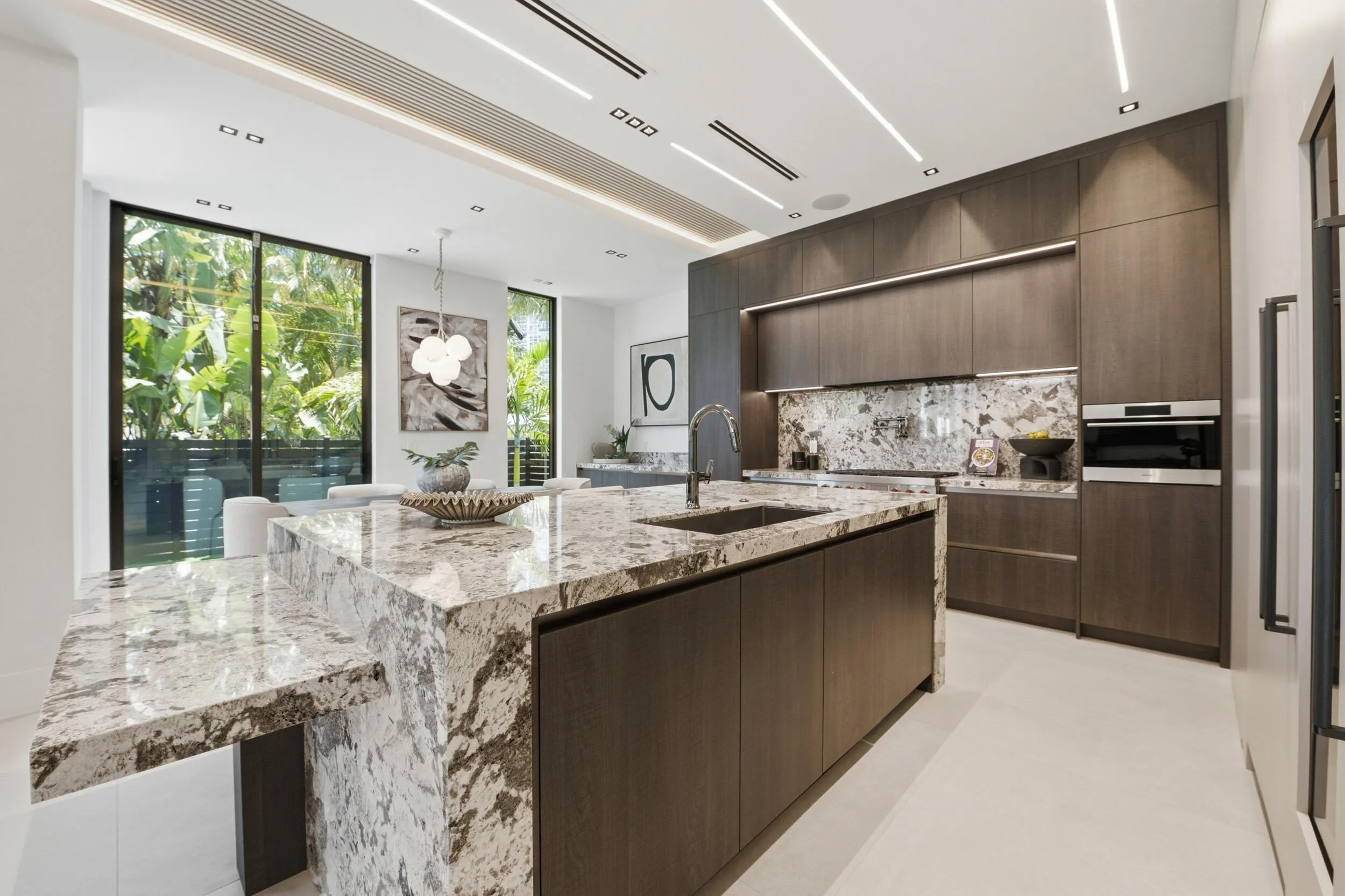 Spacious kitchen featuring a large island with an integrated breakfast bar, dark wood-finish cabinetry, built-in oven, and a complementary stone backsplash