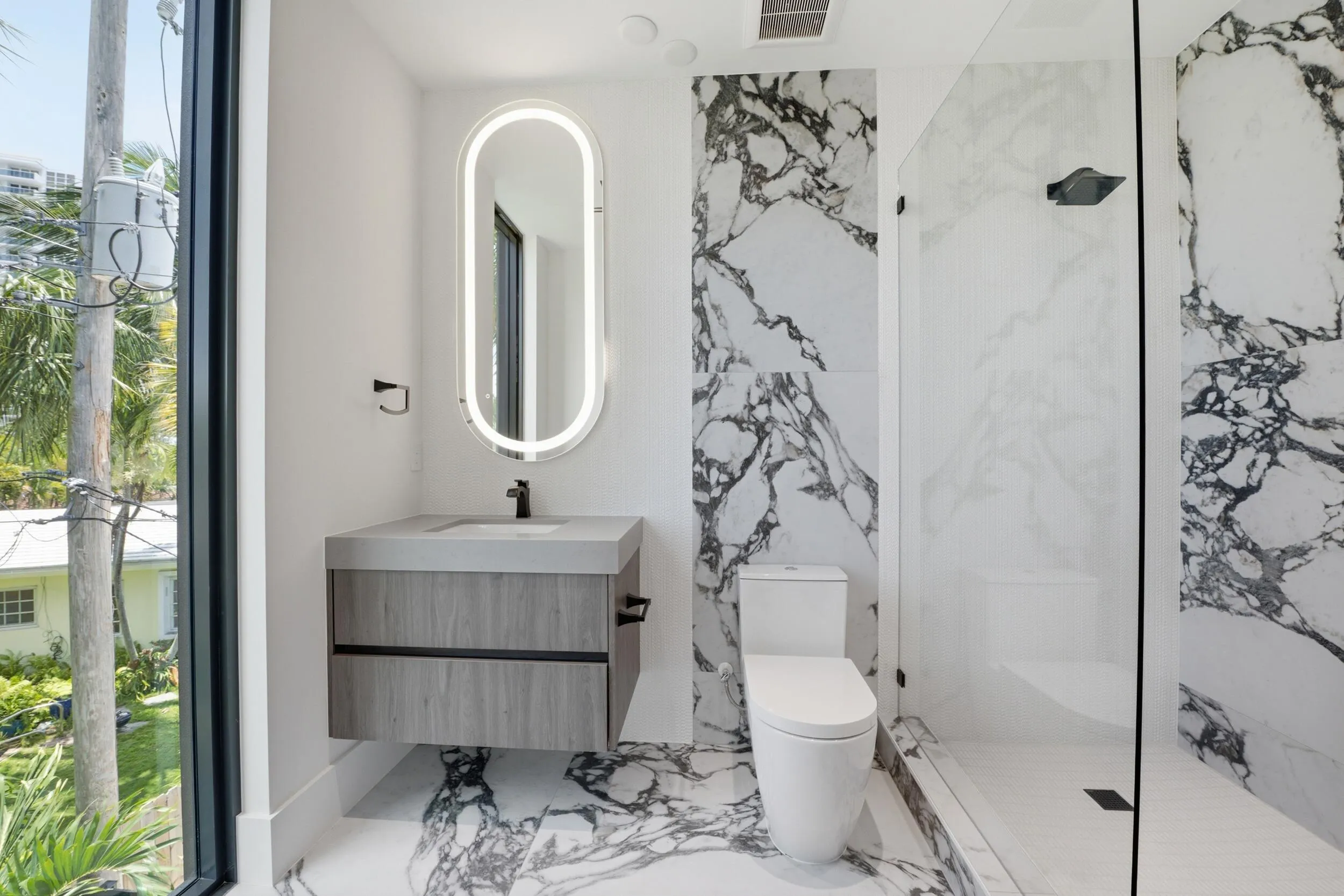 Floating vanity with integrated sink, backlit oval mirror, floor-to-ceiling tile, glass-enclosed shower, and full-height window