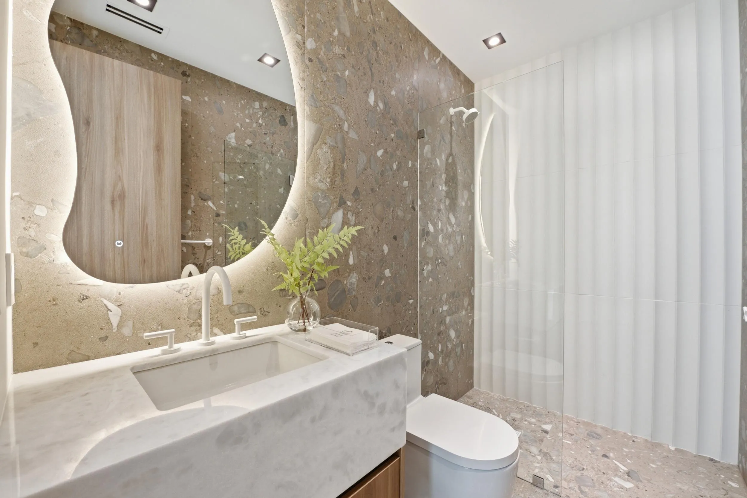 Irregularly shaped illuminated mirror, white rectangular undermount sink, three-piece white faucet set, integrated white marble countertop, and a glass-enclosed shower with fluted glass panels