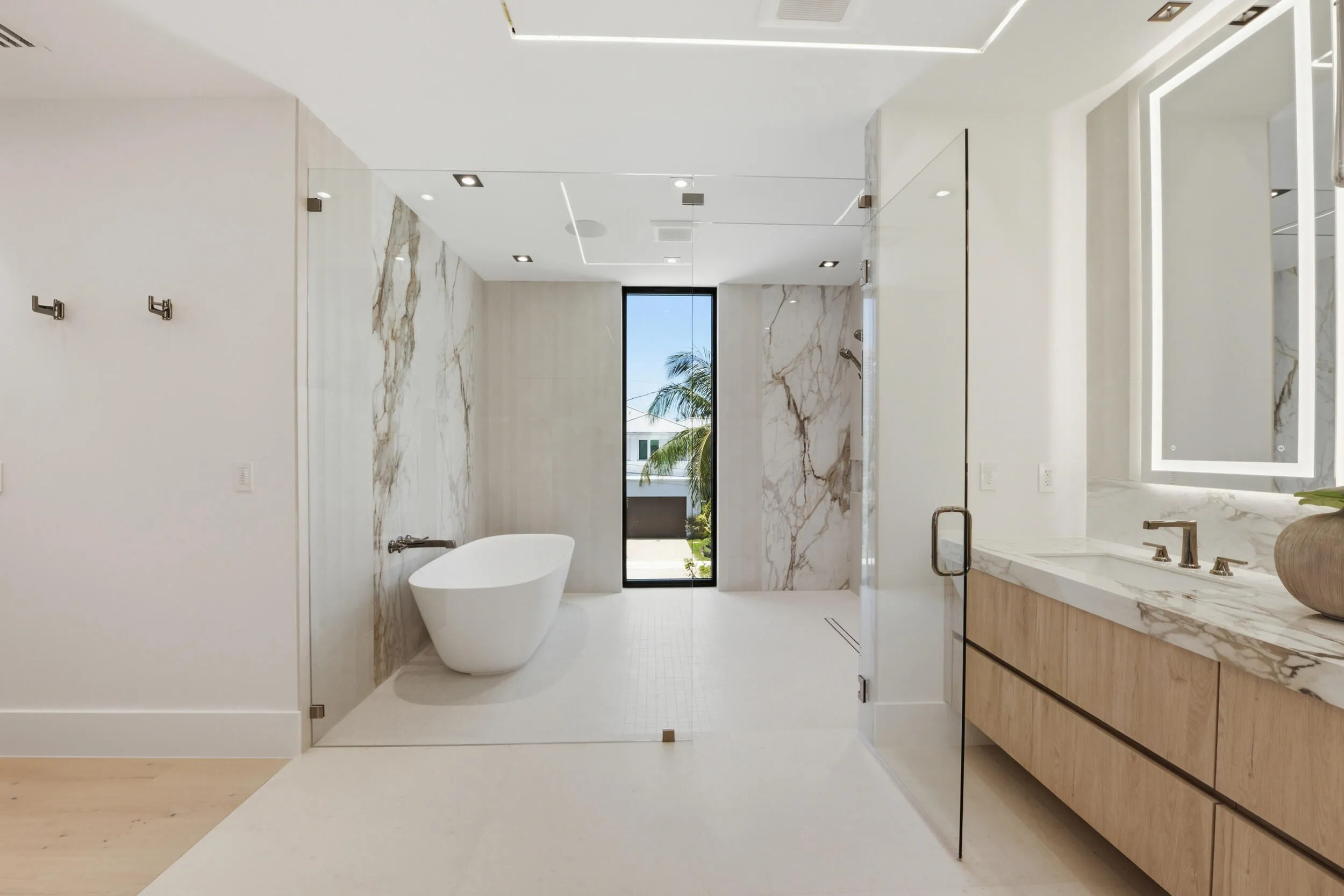 Freestanding soaking tub, floor-to-ceiling window, wood-finish vanity with stone countertop, backlit mirror, and glass-enclosed shower