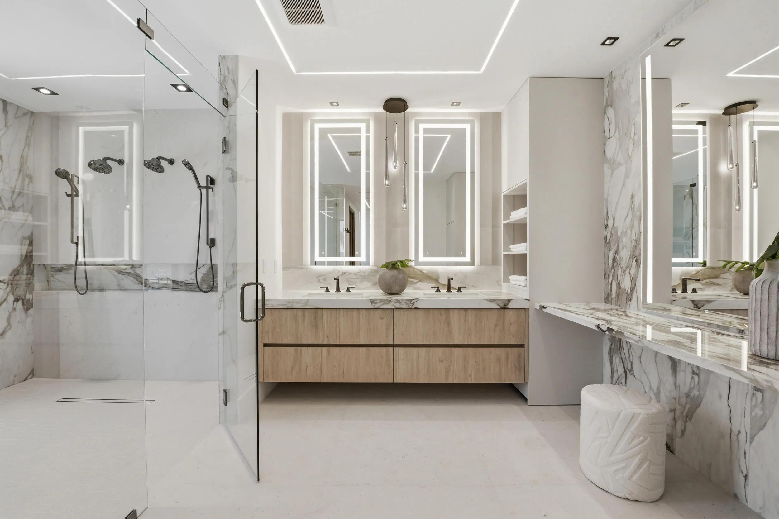 Glass-enclosed shower with multiple showerheads, dual floating vanities with integrated lighting, marble-patterned wall and vanity surfaces, and recessed ceiling lighting