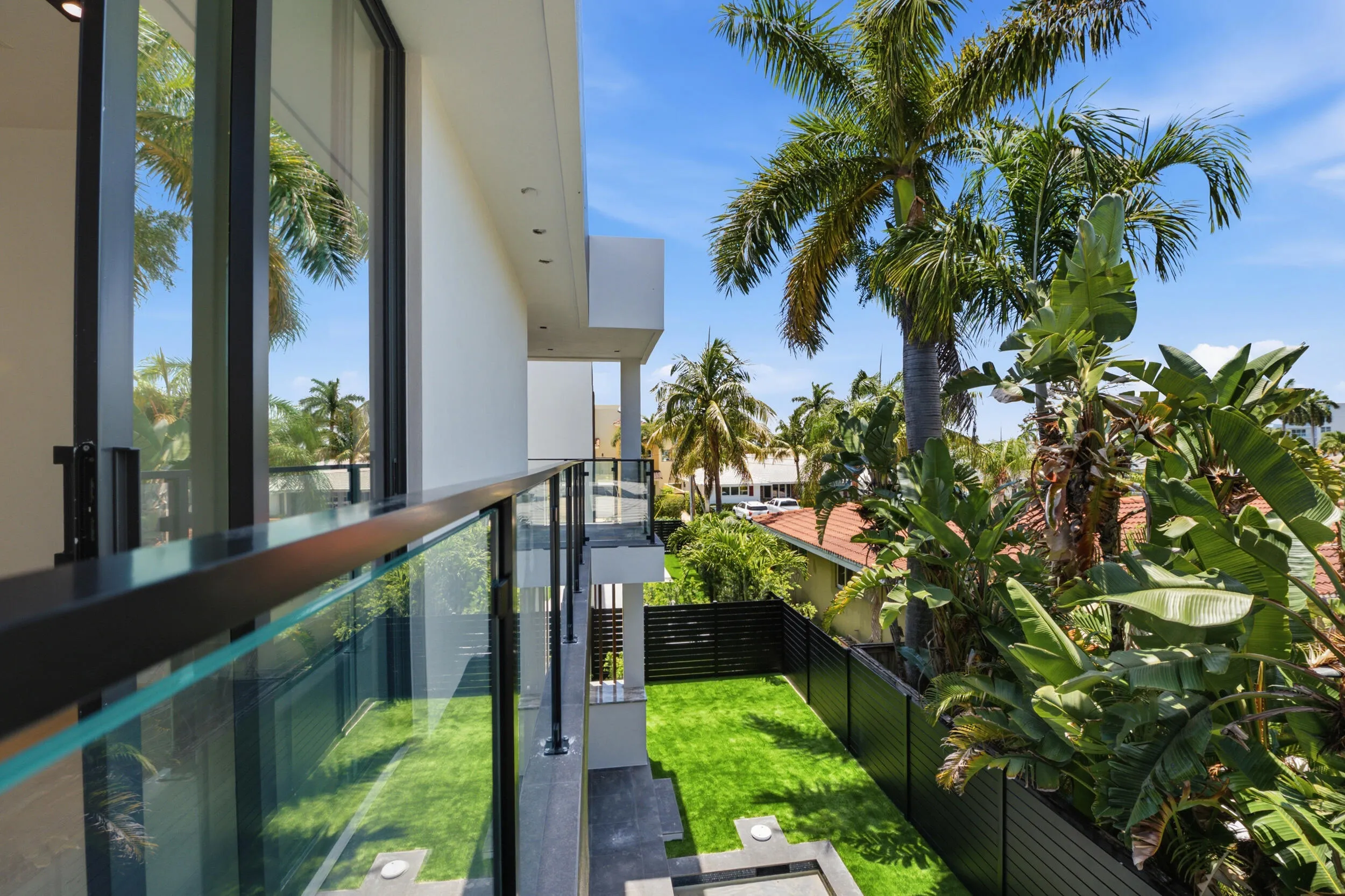 Modern glass and metal balcony railing, white exterior facade, large format sliding glass doors, private fenced yard with manicured lawn, and mature palm trees
