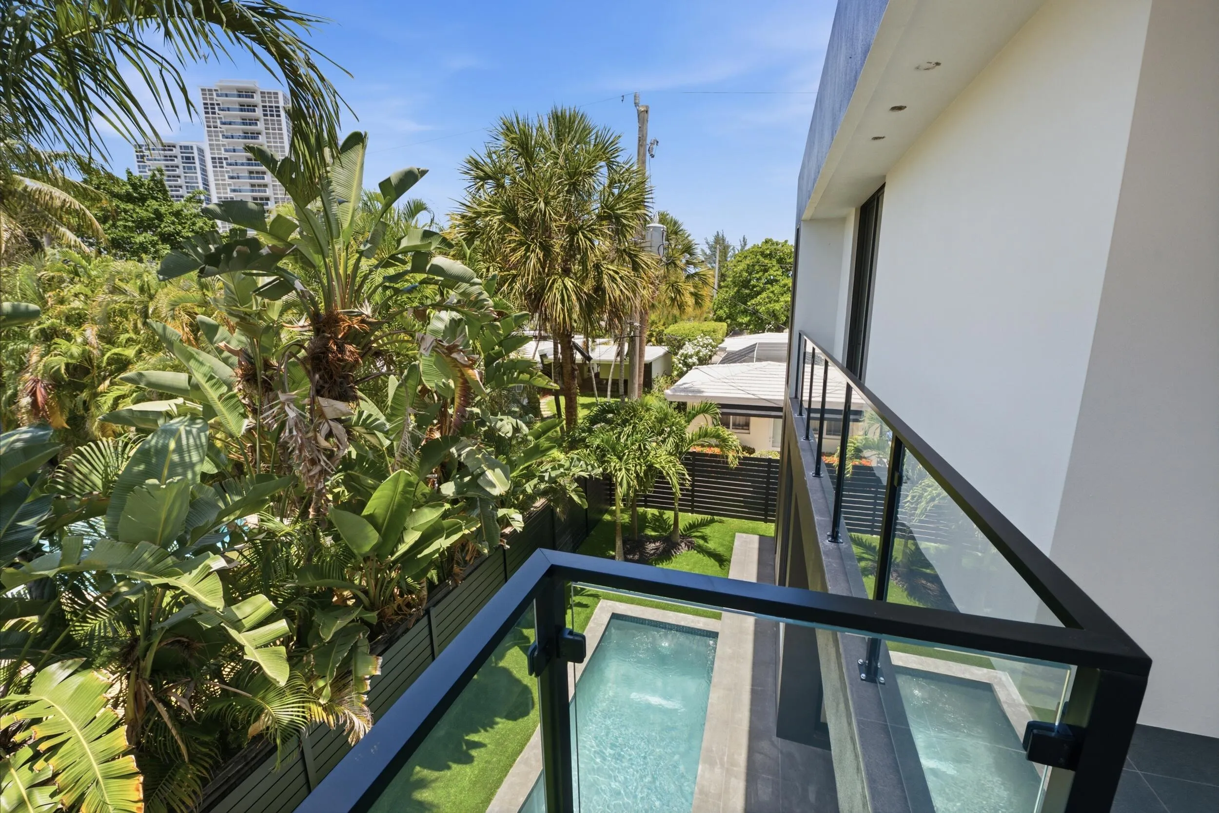 Balcony with glass panel railing and black frame, overlooking a private pool, artificial turf, and lush tropical landscaping