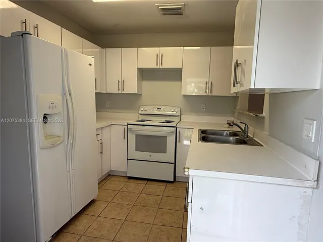 Kitchen featuring white cabinetry with metallic pulls, white appliances, solid-surface countertops, dual-basin stainless steel sink, and tile flooring