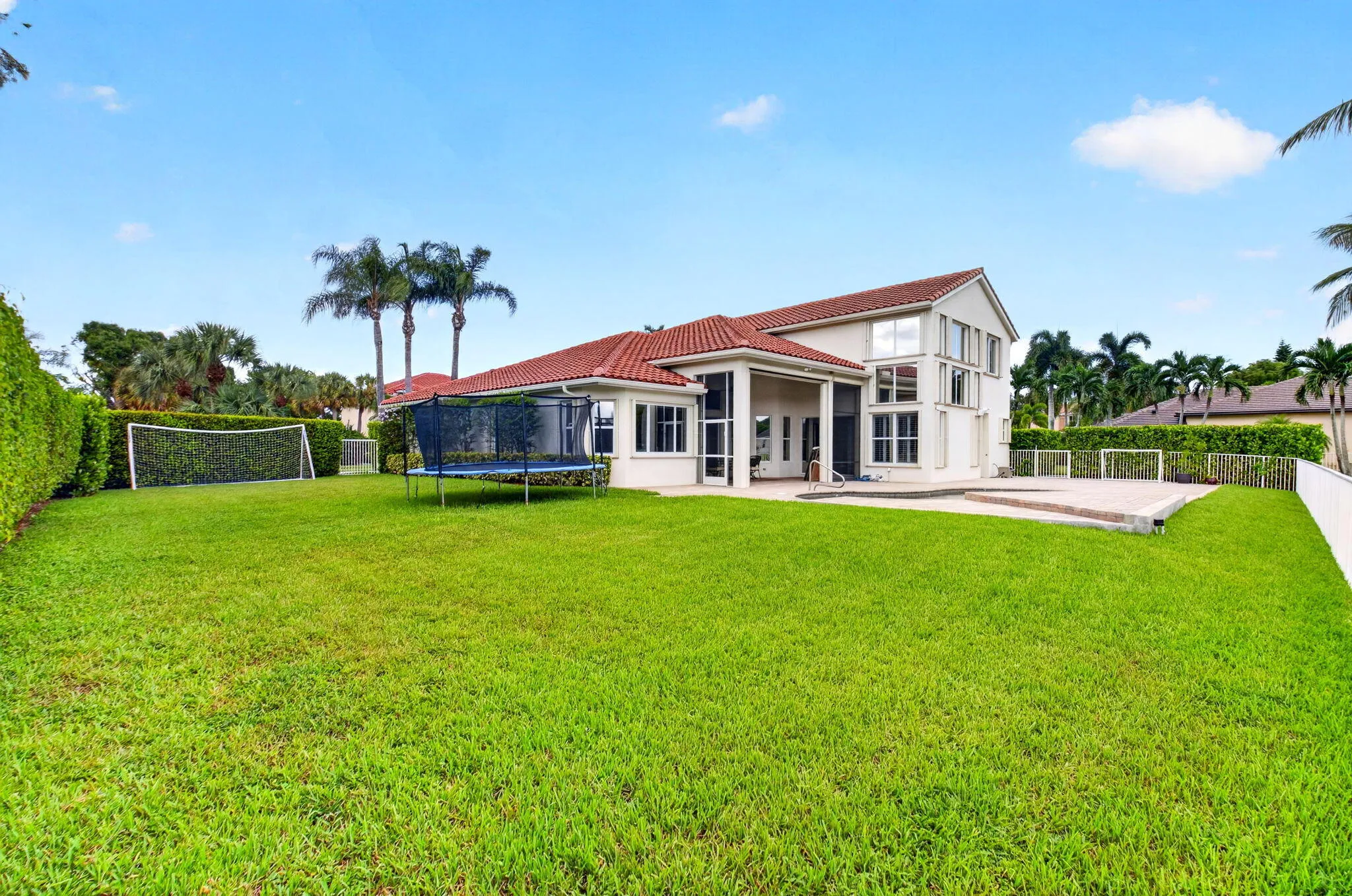 Expansive grass lawn with mature perimeter hedging, featuring a multi-story exterior with a red barrel tile roof and multiple windows, and a paved patio area