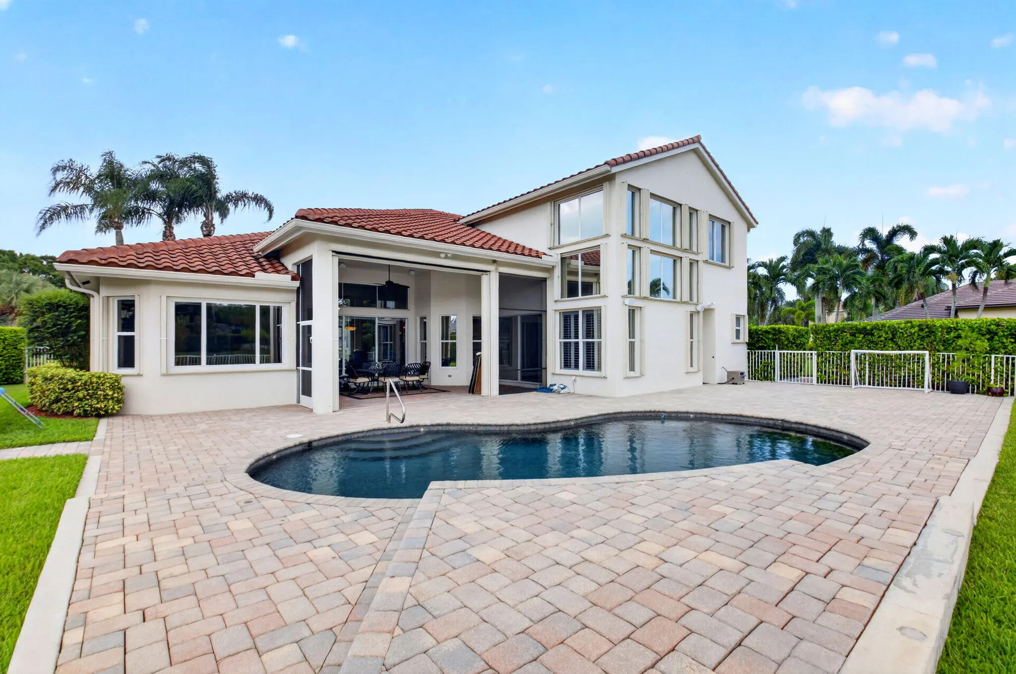Expansive paver patio surrounding a freeform swimming pool, featuring a stucco exterior with multiple large windows and a red tile roof