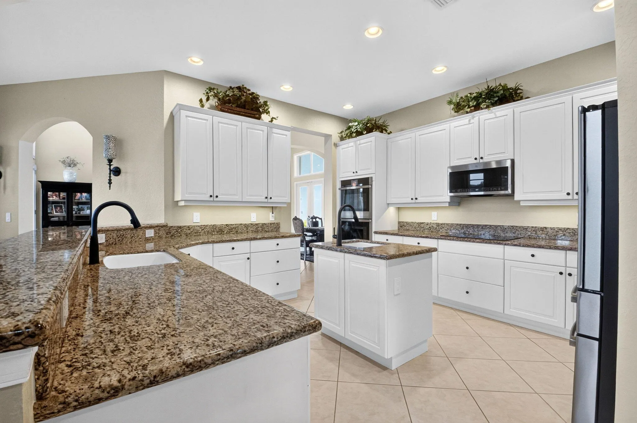 Kitchen featuring an island with an integrated sink, white cabinetry, granite countertops, and light-toned tile flooring
