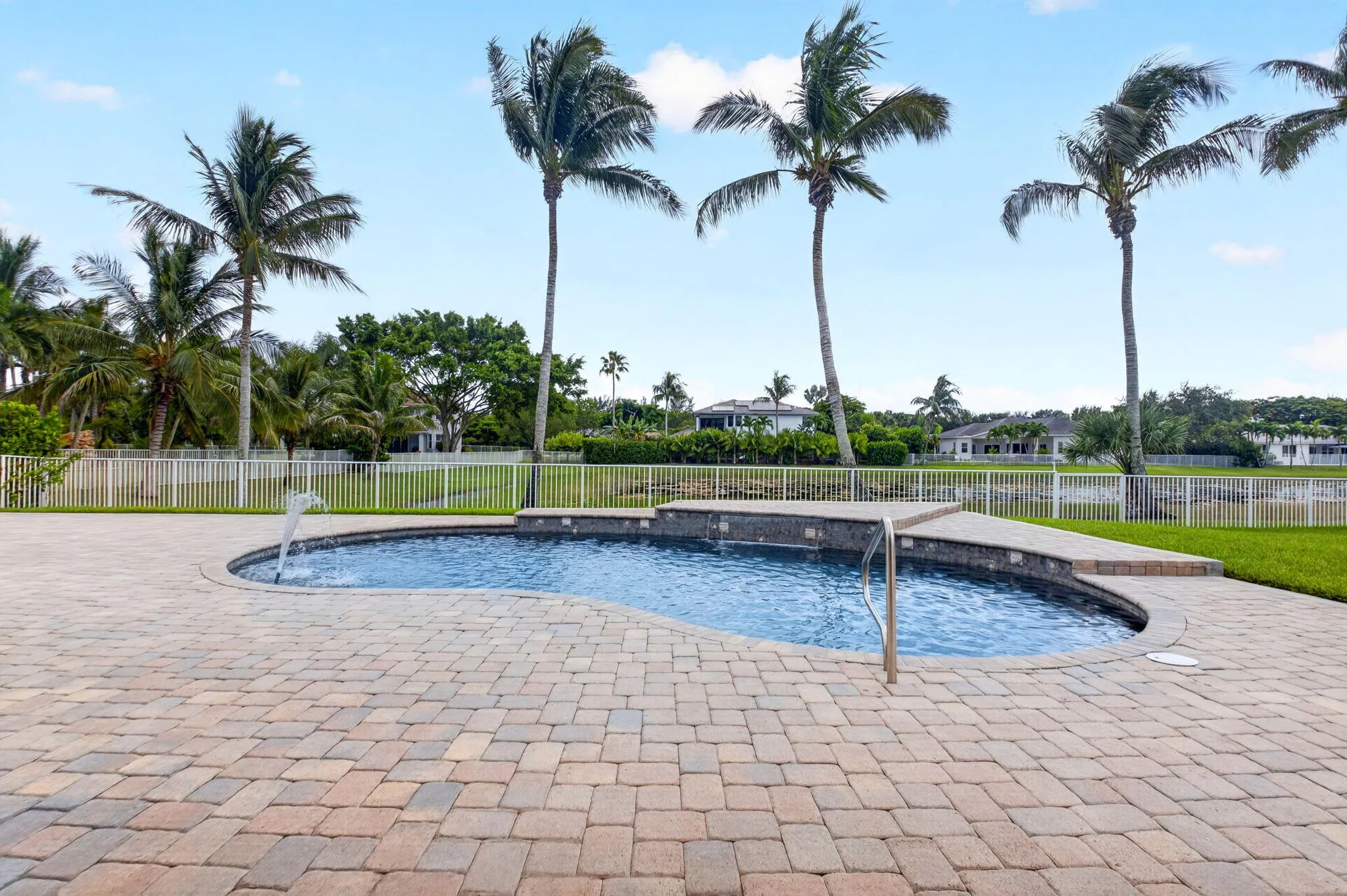 Paver patio surrounding a freeform pool with a water feature, stone tile accents, and stainless steel handrail