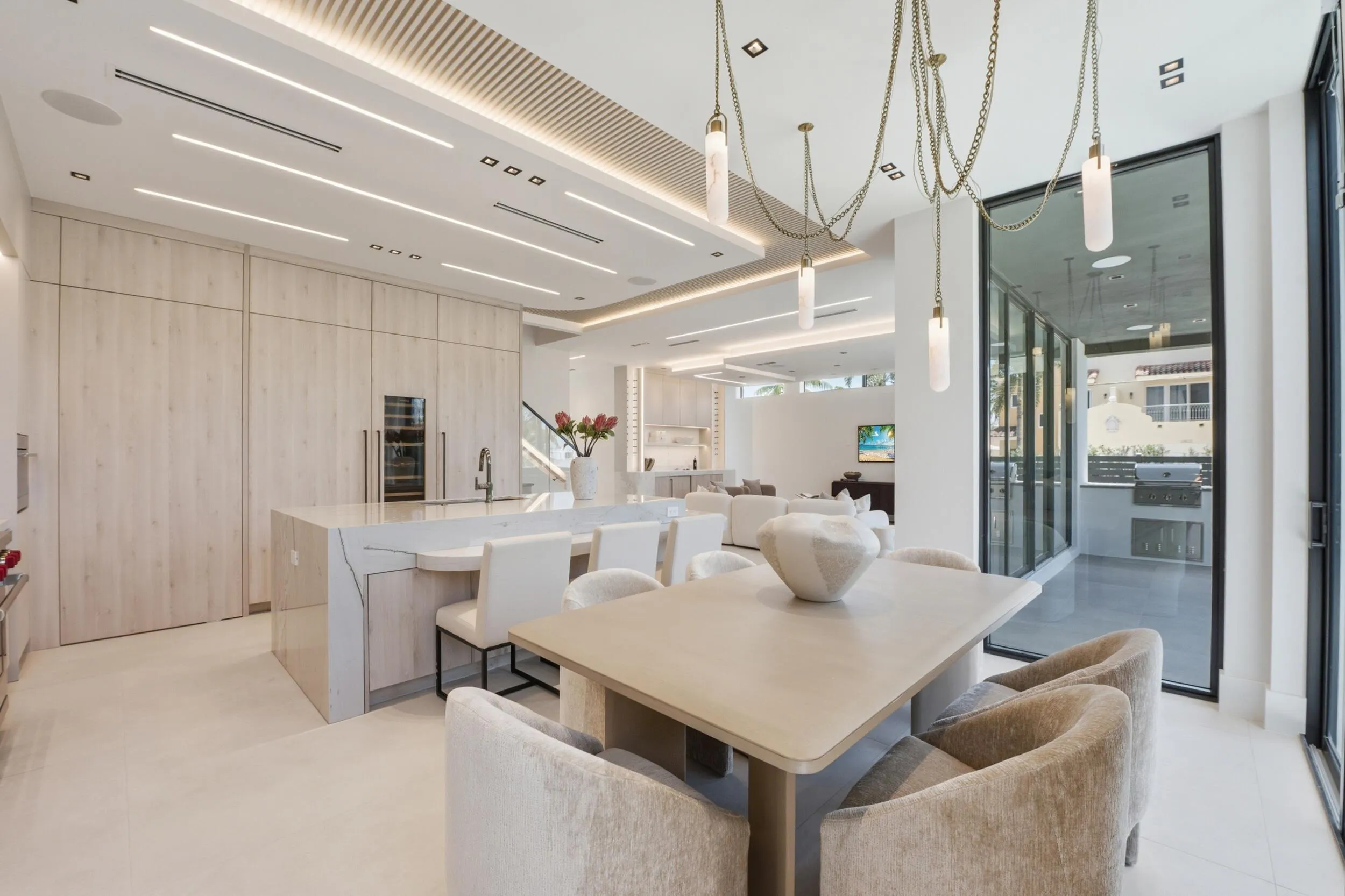 Custom kitchen cabinetry with integrated paneled appliances, large center island with waterfall countertop, recessed and linear ceiling lighting, and a multi-pendant dining chandelier