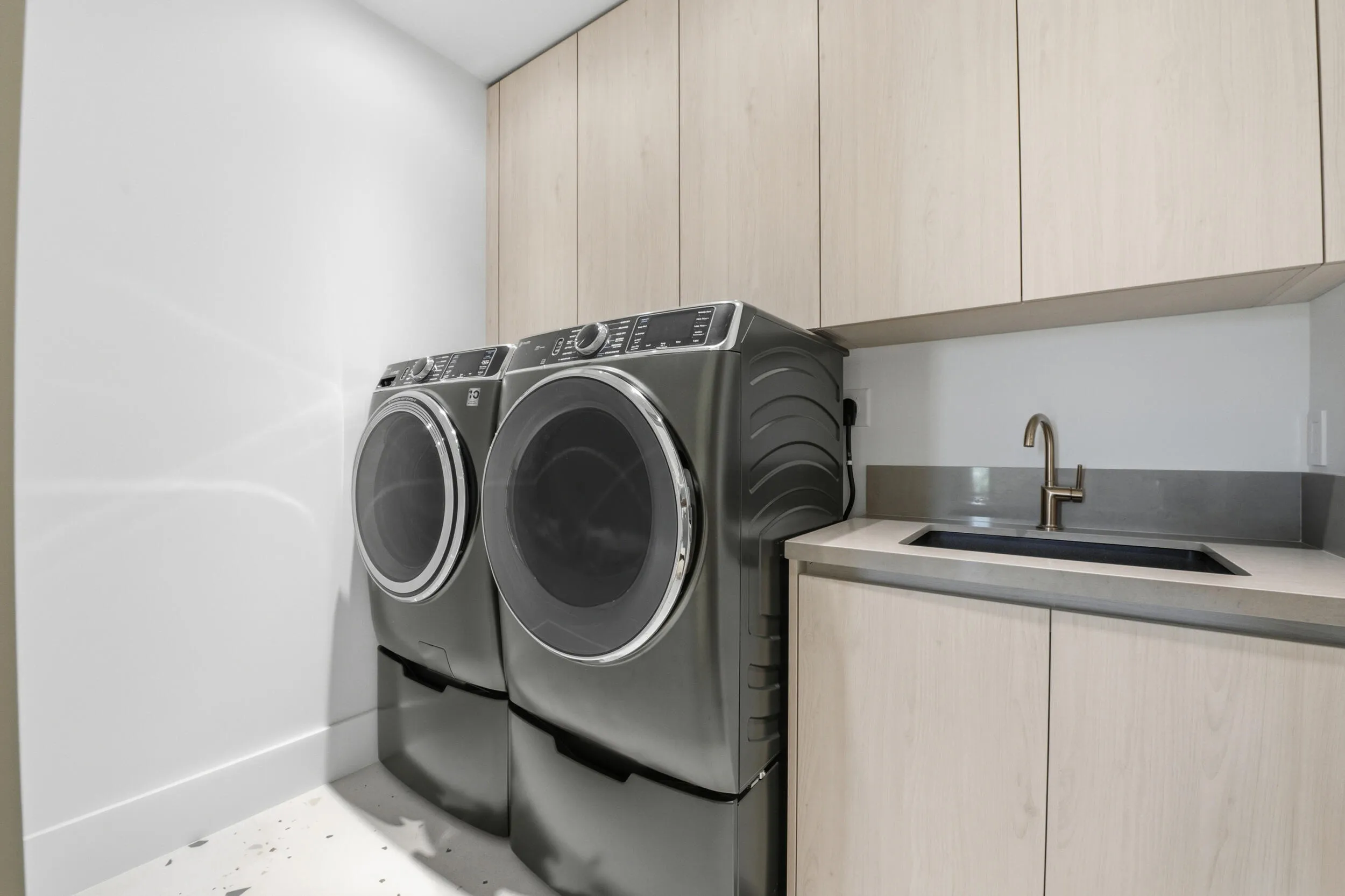 Integrated laundry area featuring light wood-finish cabinetry, a single basin sink with a bronze-tone faucet, and a grey countertop