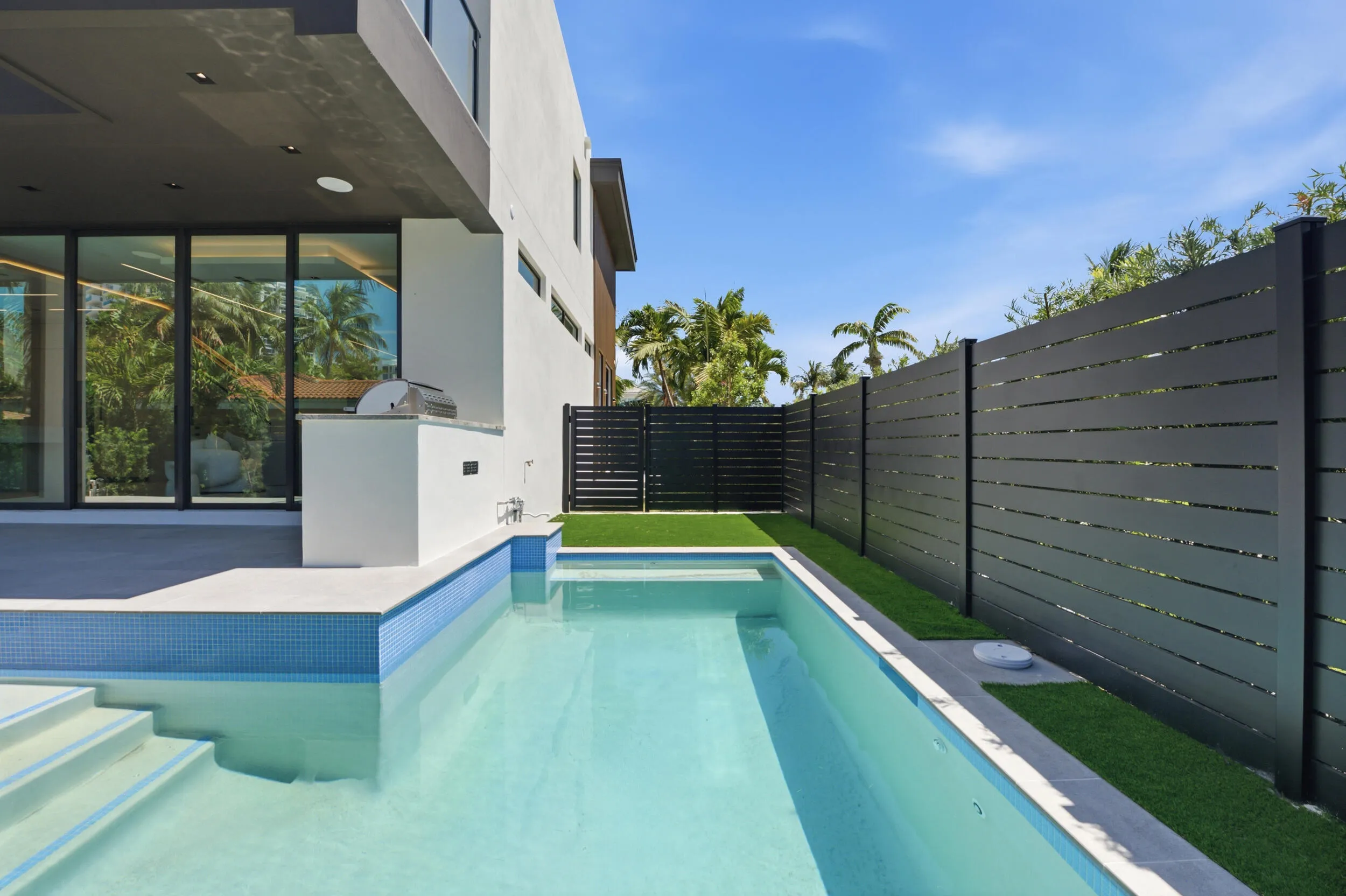 Contemporary exterior featuring a rectangular pool, integrated spa, built-in outdoor kitchen with grill, expansive sliding glass doors, and a privacy fence