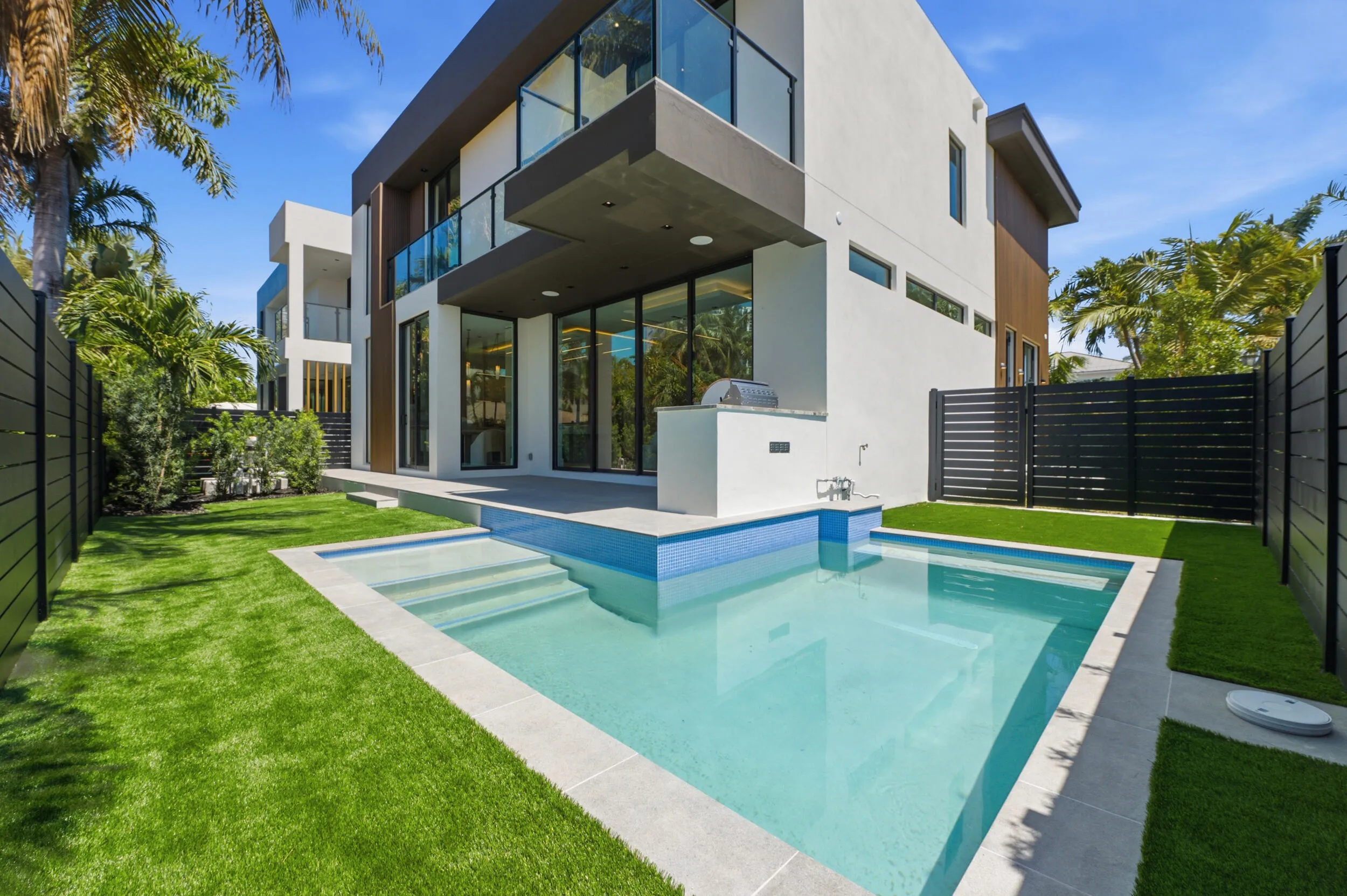 Contemporary exterior with a private swimming pool, built-in outdoor kitchen, expansive glass doors, second-story balcony with glass railing, and a manicured lawn