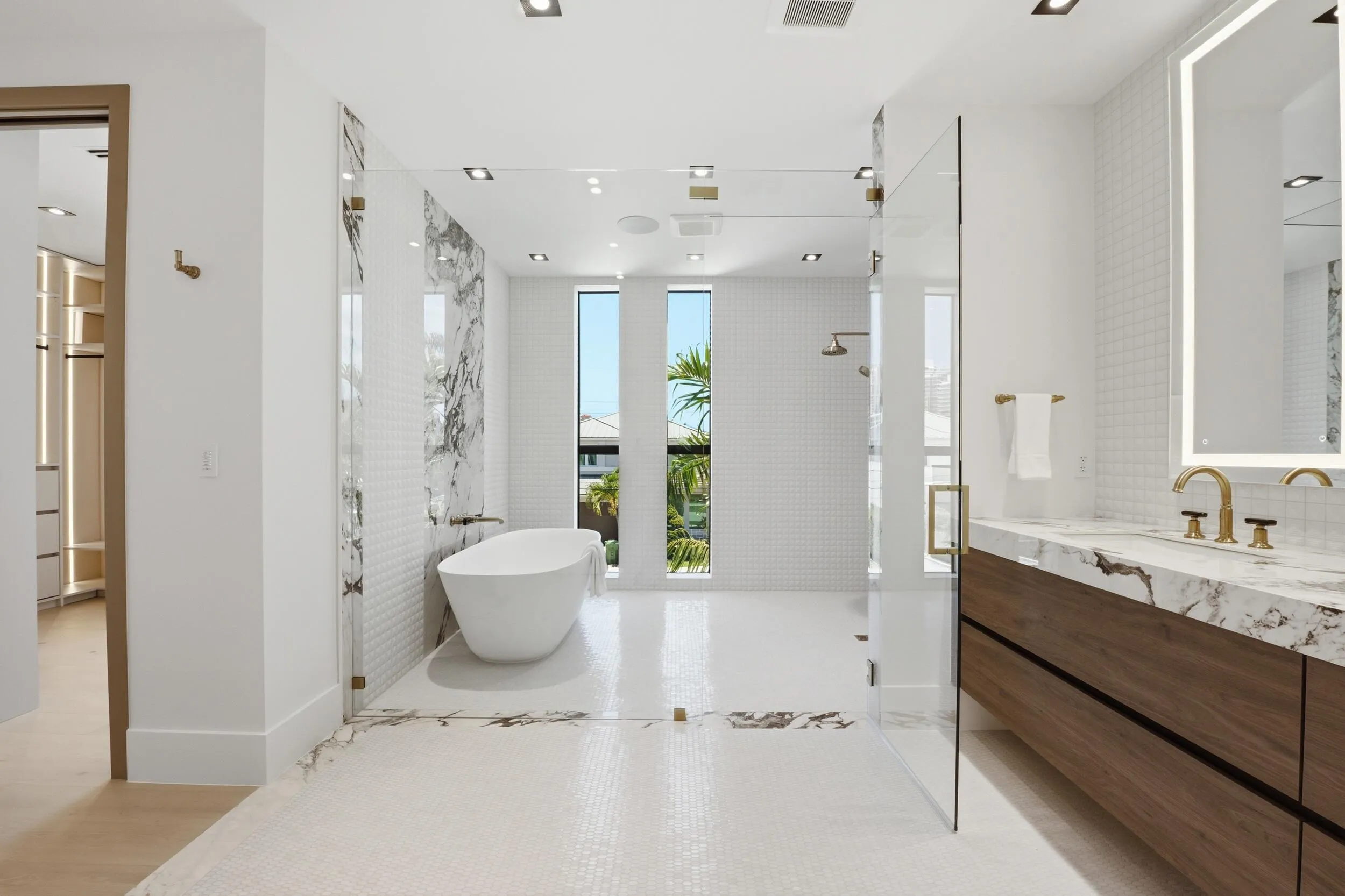 Freestanding soaking tub, glass-enclosed shower, dual vanity with wood-finish cabinetry, marble-look countertops, and gold-tone fixtures