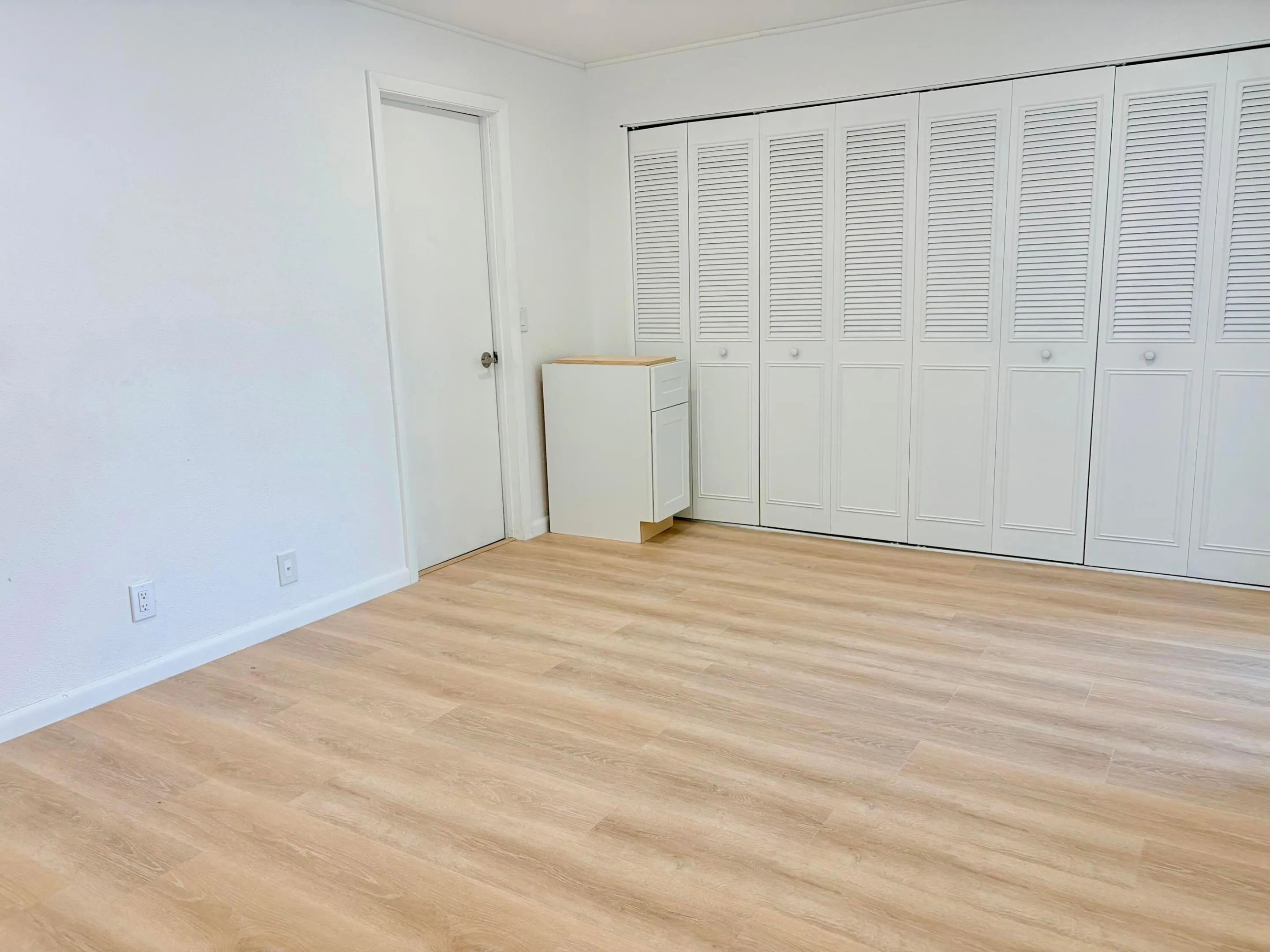 Wood-finish flooring, white walls, white louvered bifold closet doors, white interior door, and a light-toned wood-finish counter