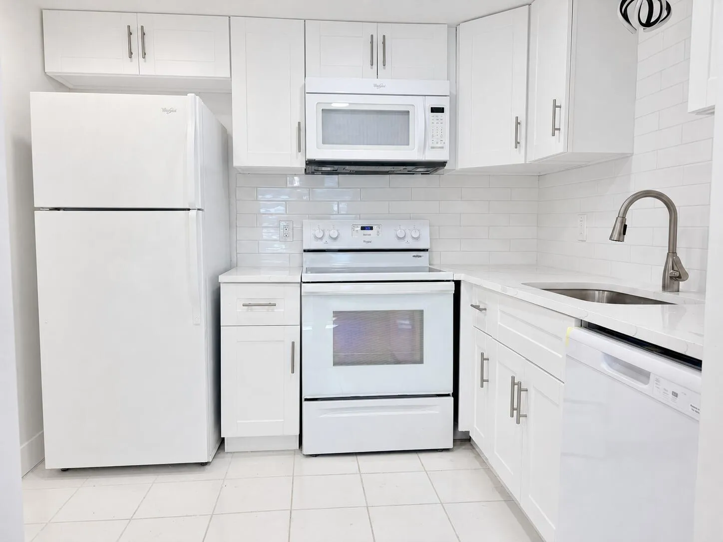 White shaker-style cabinetry with minimalist hardware, white subway tile backsplash, stainless steel sink with gooseneck faucet, white appliance suite including dishwasher and microwave, and light-toned flooring
