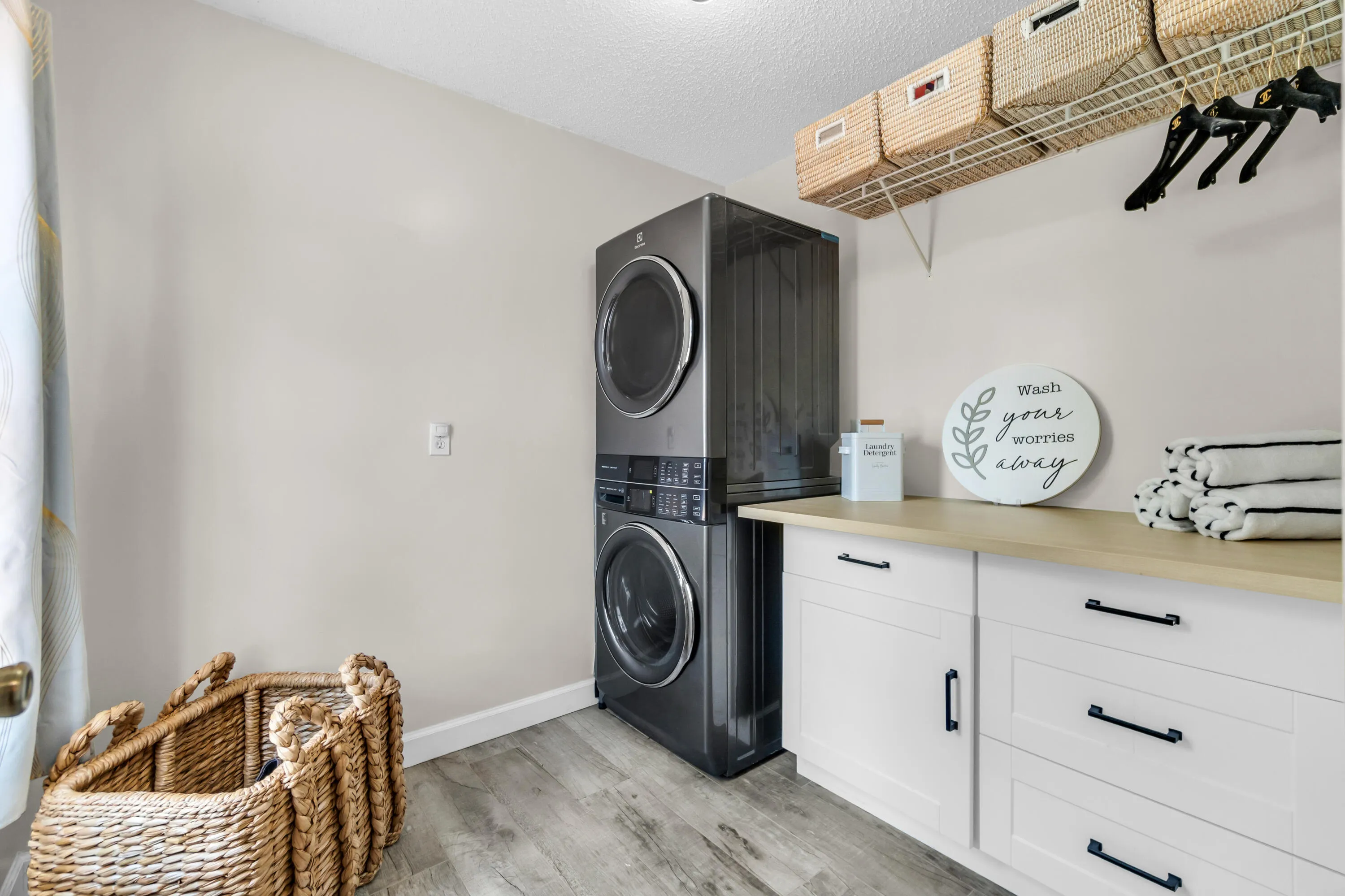 Laundry area with wood-finish flooring, stacked washer and dryer, built-in white cabinetry with black hardware, wood-finish countertop, and overhead wire shelving