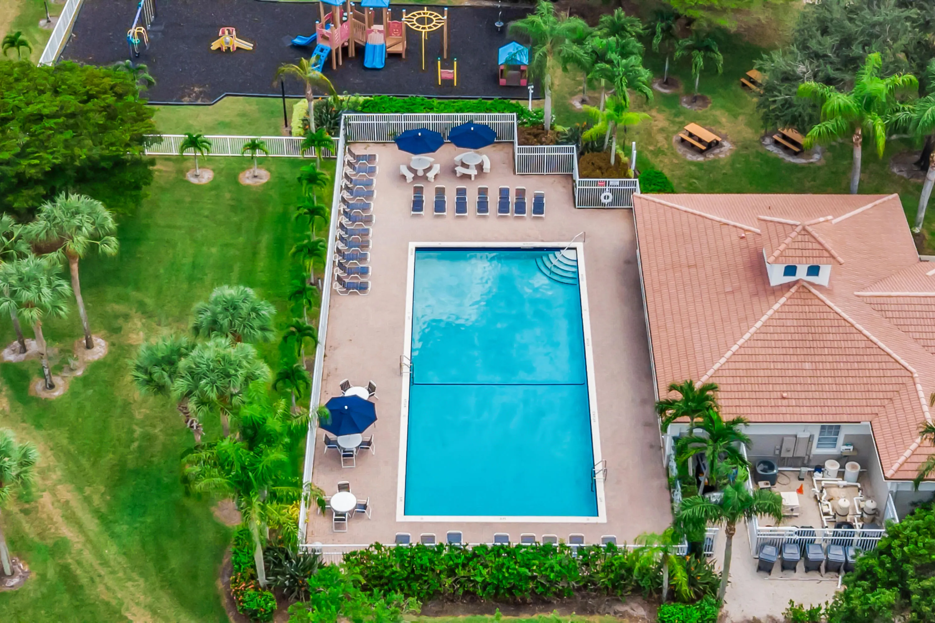 Community swimming pool with surrounding lounge seating, adjacent clubhouse with a tile roof, expansive green spaces, and a nearby recreational play area