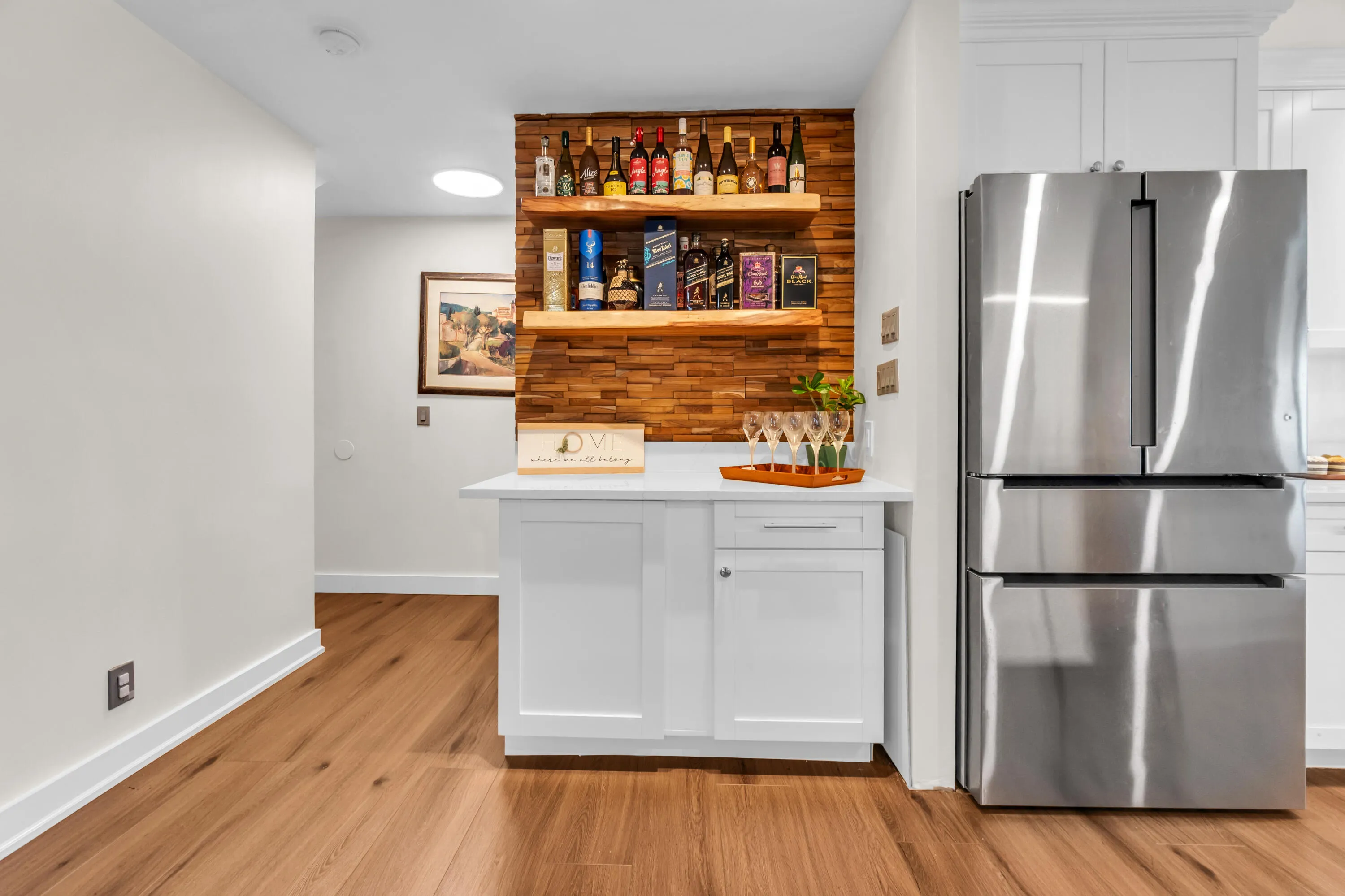 White cabinetry with a light-toned countertop, wood-finish flooring, a stainless steel refrigerator, and a wood-paneled accent wall with floating shelves
