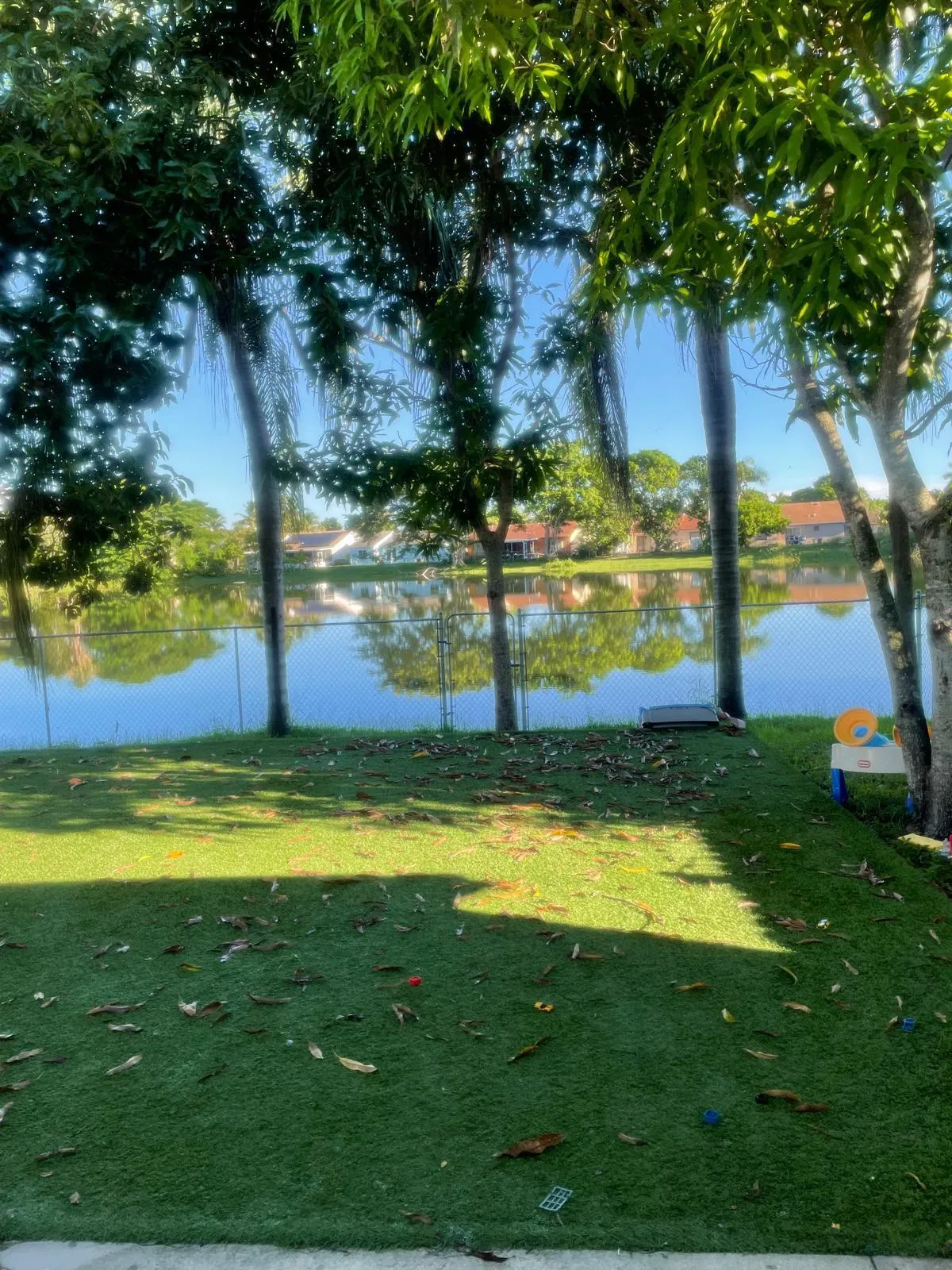 Fenced backyard featuring an artificial turf lawn, mature trees, and a tranquil water feature